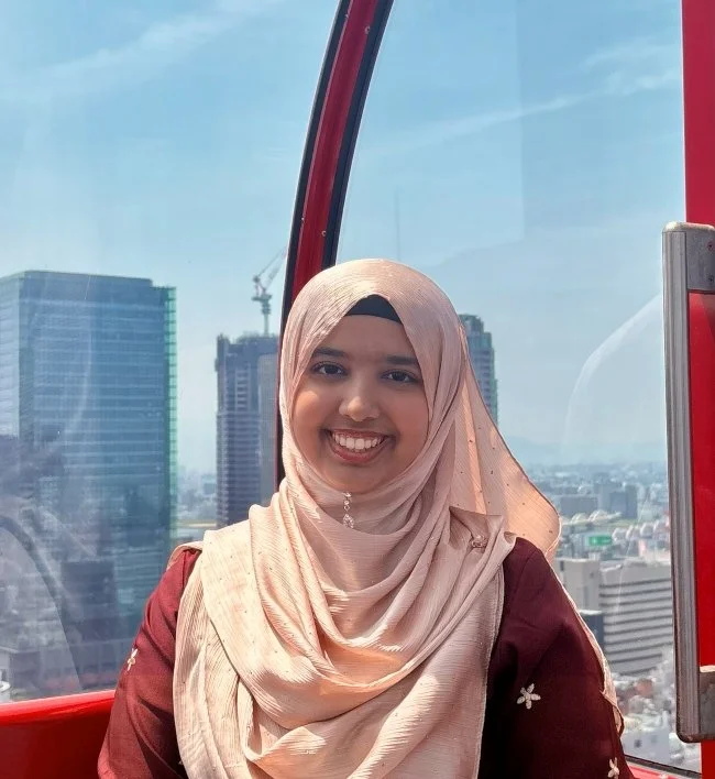 A young woman with a beige hijab and a maroon top smiling inside a red gondola cabin with a city skyline in the background.
