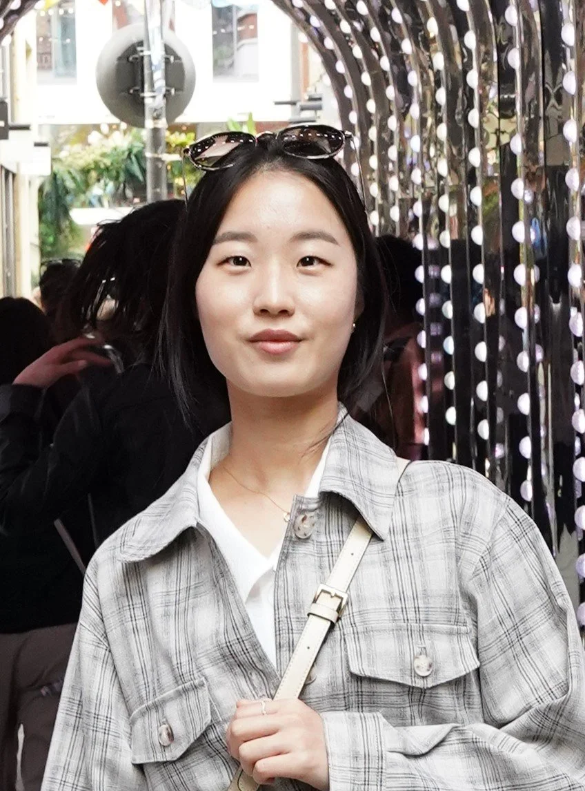 A young woman with short black hair and light skin standing outdoors in front of a decorative light installation. She is wearing a plaid jacket, white shirt, and sunglasses on her head, and carrying a beige shoulder bag.