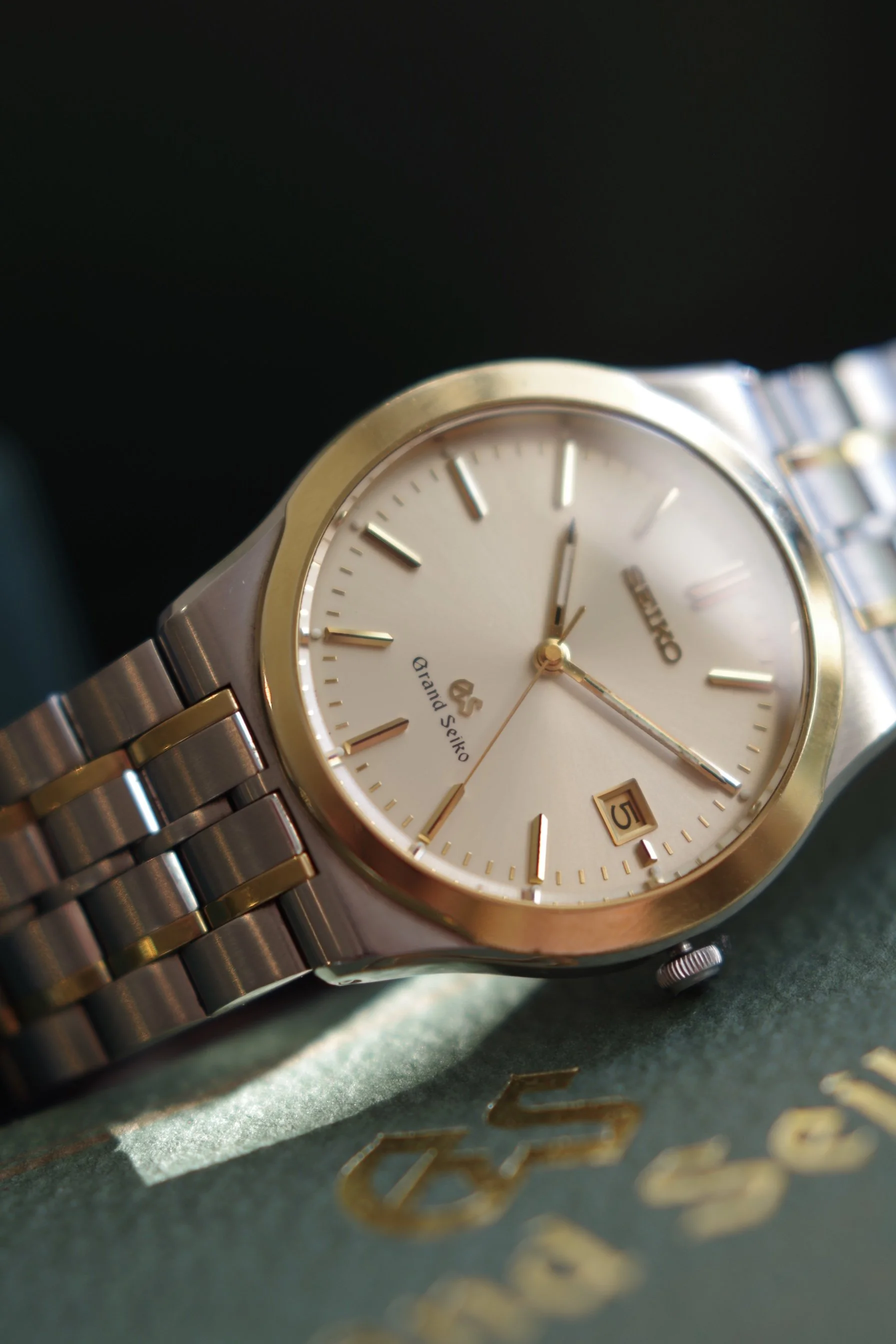 A Grand Seiko wristwatch with a silver and gold two-tone bracelet, a white dial, gold hour markers, and a date window at 4:30. The watch is resting on a green surface with gold lettering.