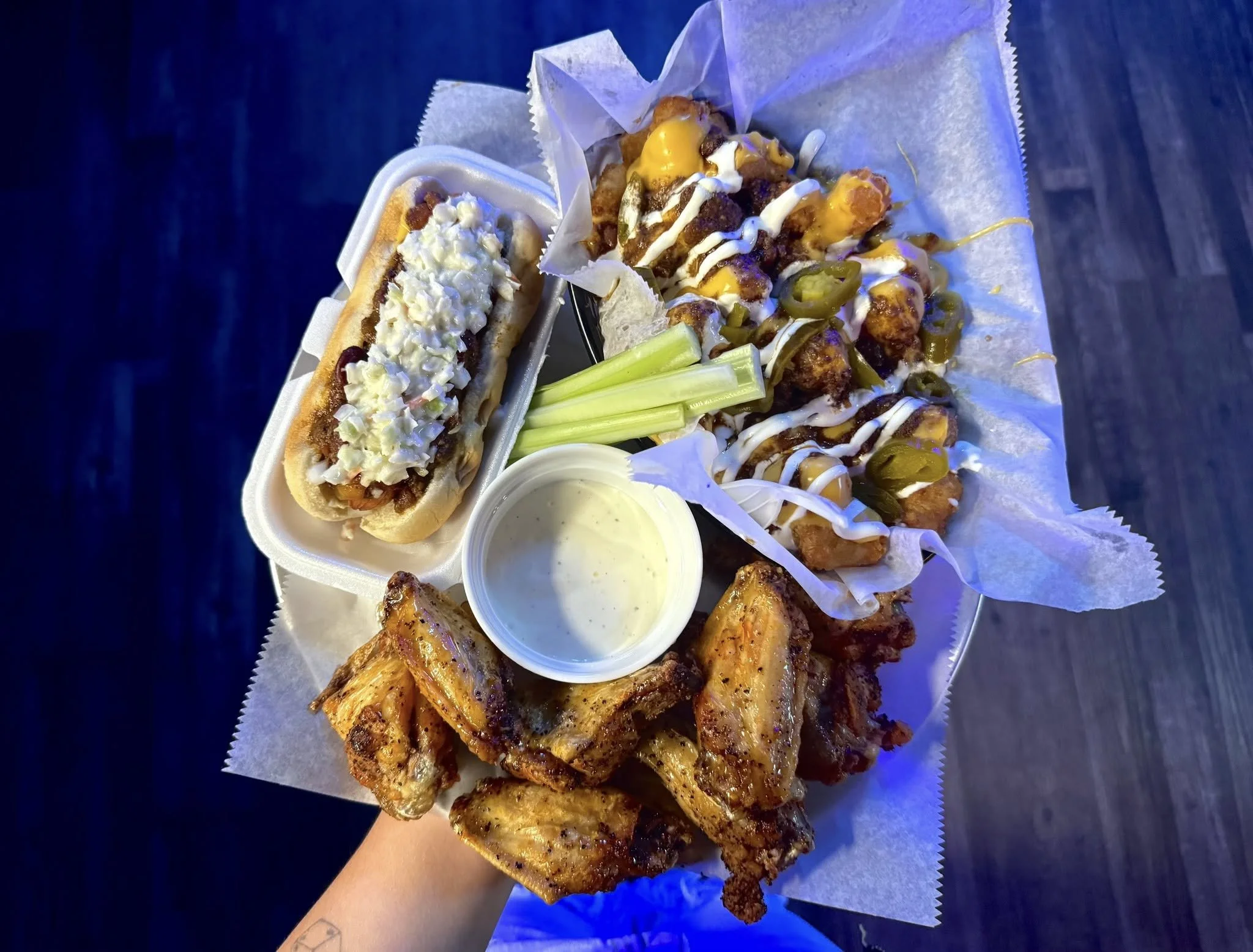A tray holding a hot dog with toppings, buffalo chicken wings with dipping sauce, fried chicken bites with sauces, celery sticks, and a cup of ranch dressing.