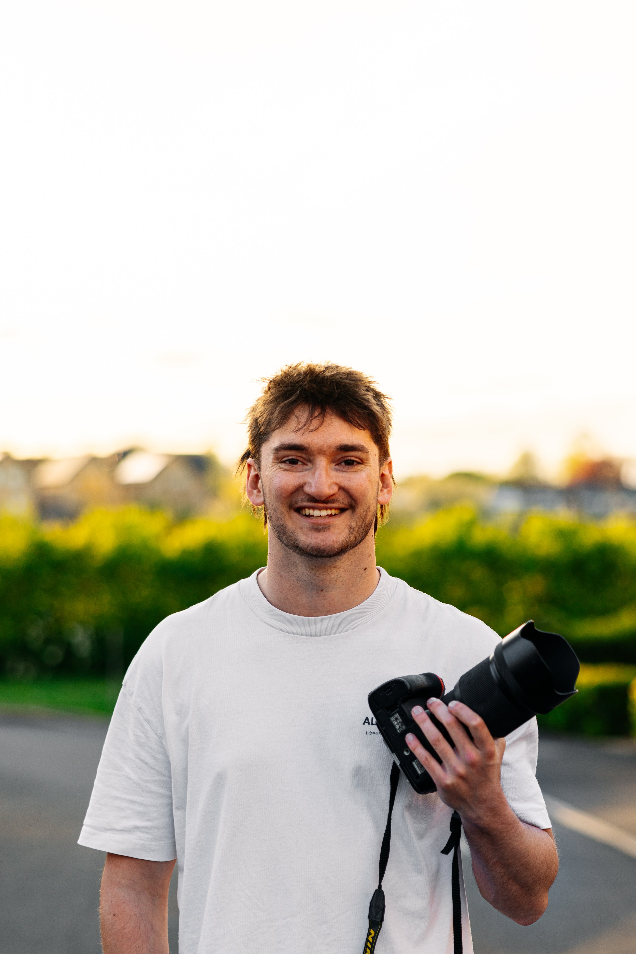 A young man smiling outdoors, holding a camera with a zoom lens, wearing a white T-shirt and standing in a green outdoor setting during sunset.