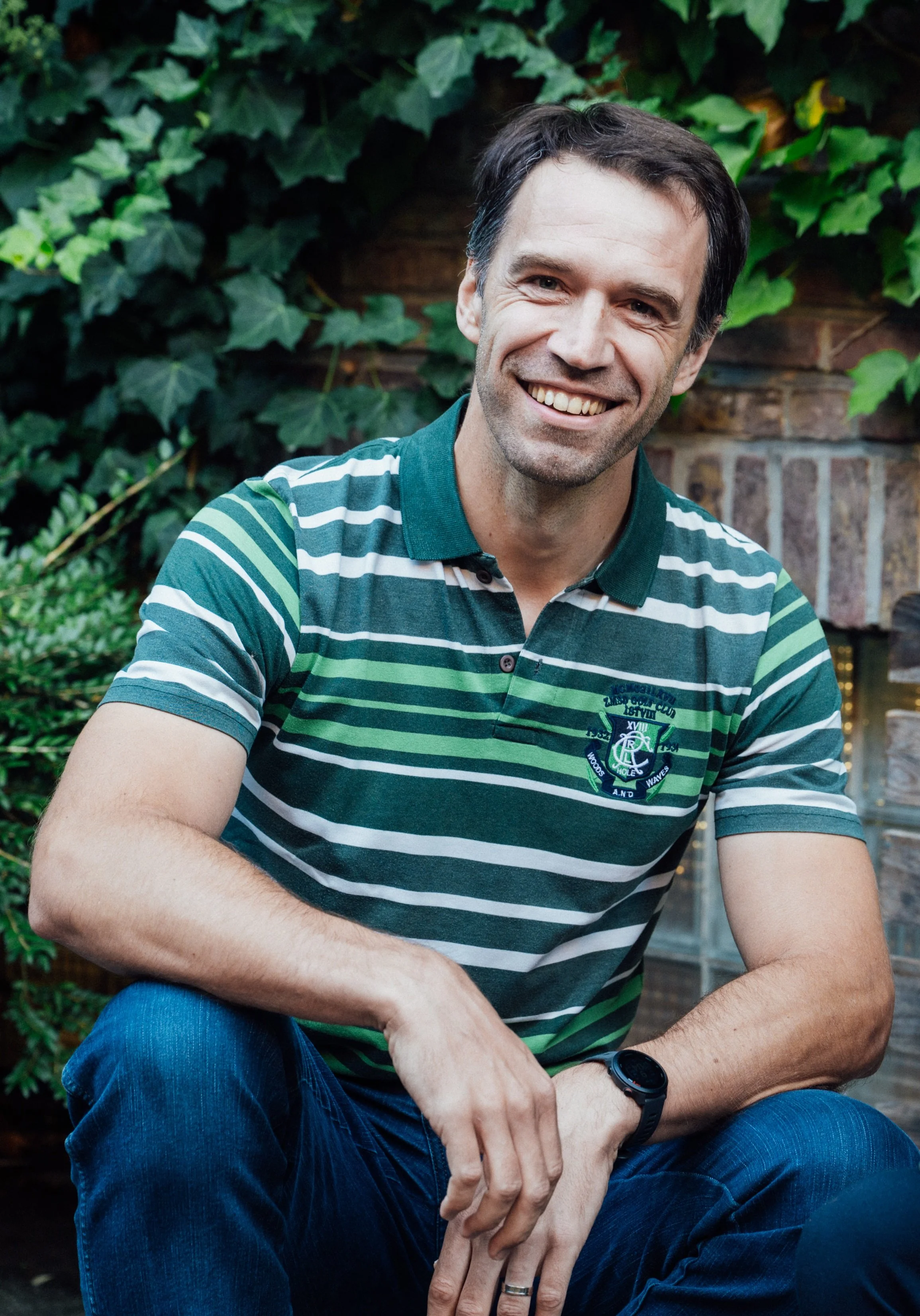 A smiling man with dark hair and a five o'clock shadow, wearing a green and white striped polo shirt, sitting outdoors with greenery and brick wall in the background.