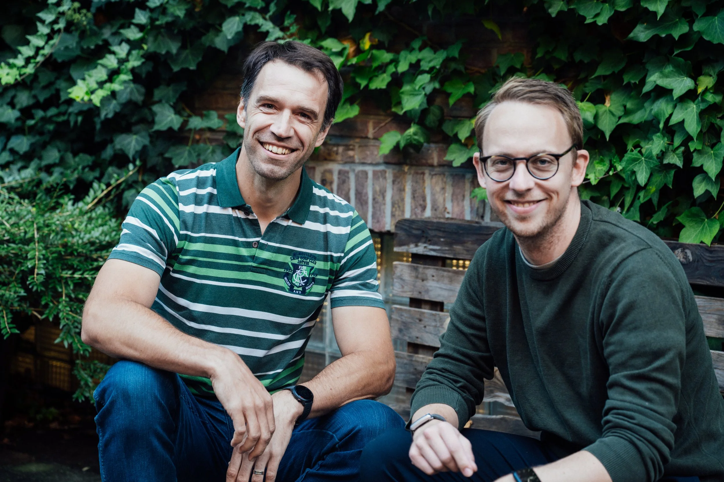 Two smiling men sitting outdoors on a wooden bench with greenery and ivy in the background.