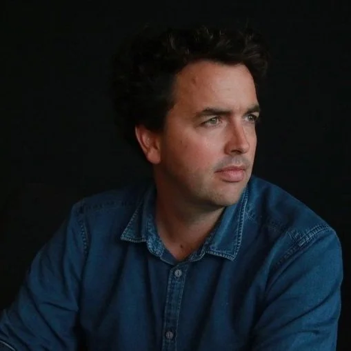 A man with dark, curly hair and light skin wearing a blue denim shirt, looking to his right against a black background.
