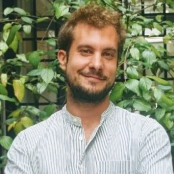 A man with curly hair and a beard smiling, wearing a striped shirt, standing outdoors in front of green plants.