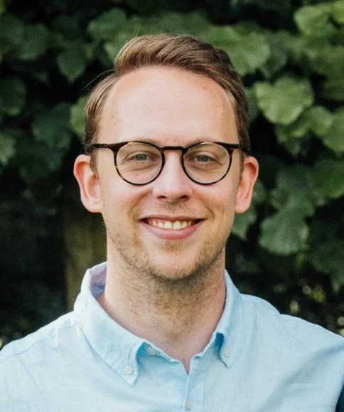 A smiling young man with glasses and light brown hair, wearing a light blue button-up shirt, standing outdoors with green foliage in the background.