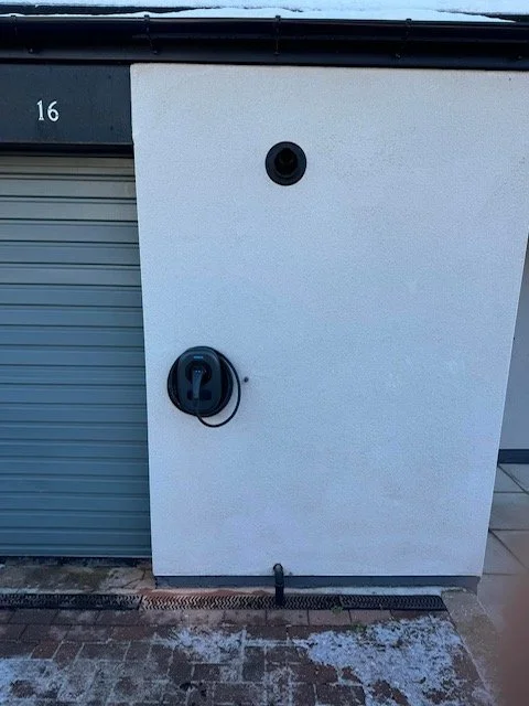 White utility box with a black security camera and an electrical outlet with a cable in front of a building with a garage door.