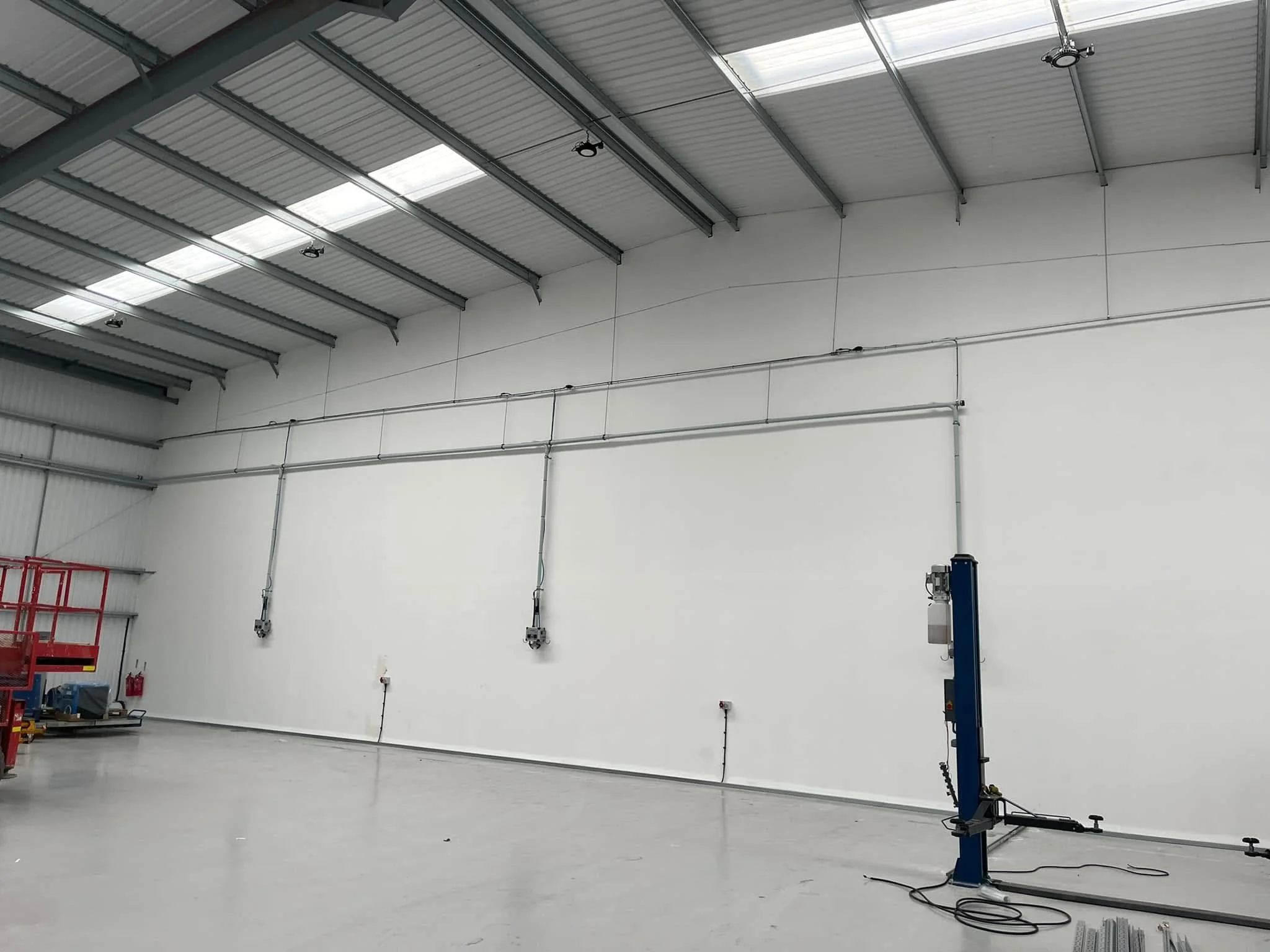 Interior of an empty industrial warehouse with high metal ceiling, white walls, and concrete floor; some equipment and electrical installations along the walls.