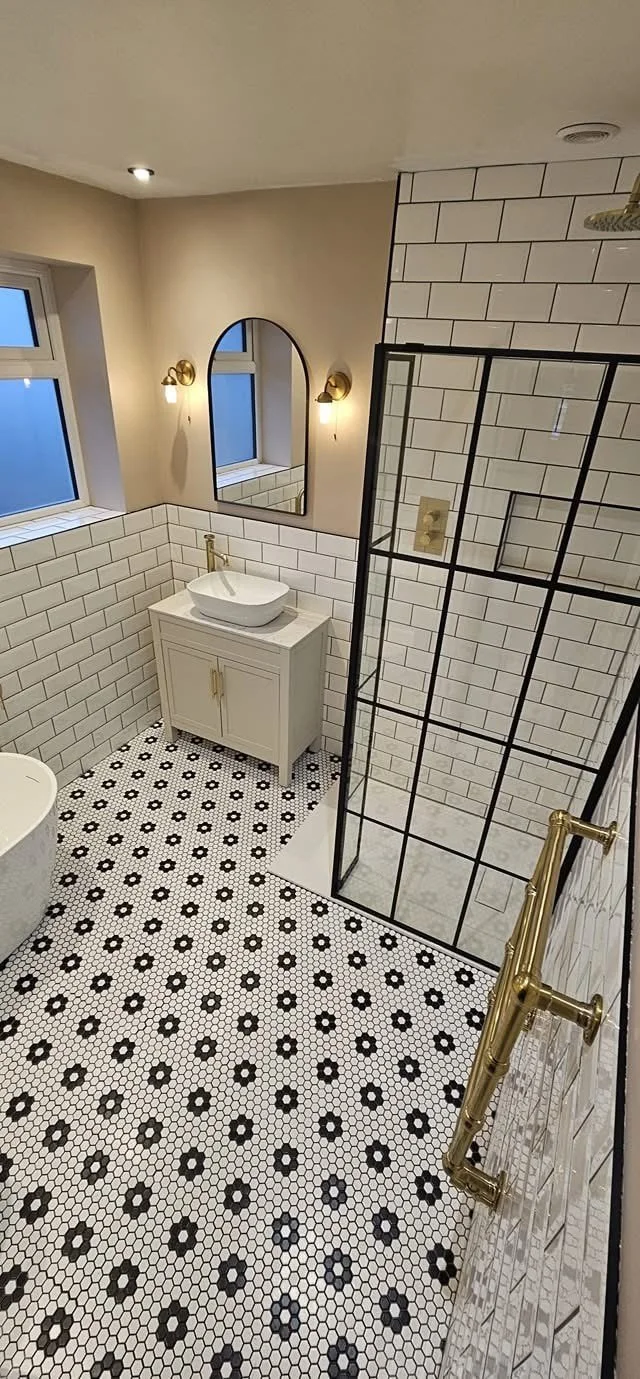 A modern bathroom with white subway tile walls, a black-framed glass shower, patterned black and white tile floor, a white vanity with a vessel sink, a large mirror, and gold fixtures, illuminated by wall sconces and ceiling lights.