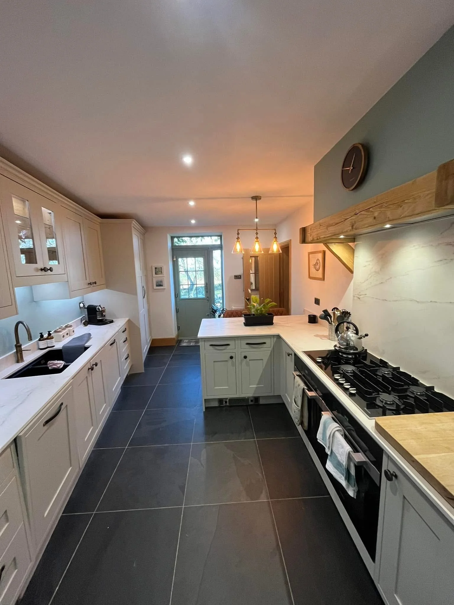 Modern kitchen with white cabinets, black tiled floor, and a wooden range hood. Contains a stove, kettle, potted plant, and hanging lights. At the back, there is a door with windows leading outside.