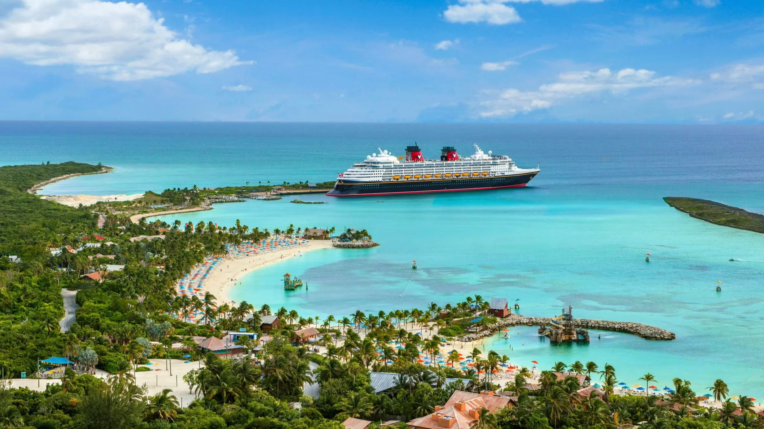 Large cruise ship anchored near a tropical island with a sandy beach, palm trees, and colorful umbrellas, under a partly cloudy blue sky.
