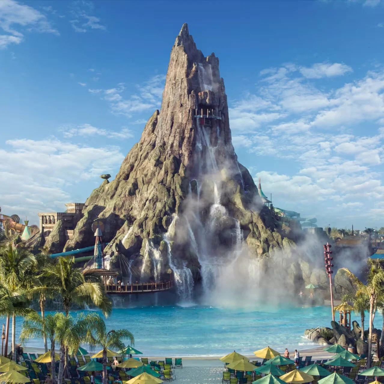 A large artificial volcano with water cascading down its sides, surrounded by a water park with palm trees, umbrellas, lounge chairs, and people enjoying the scene.