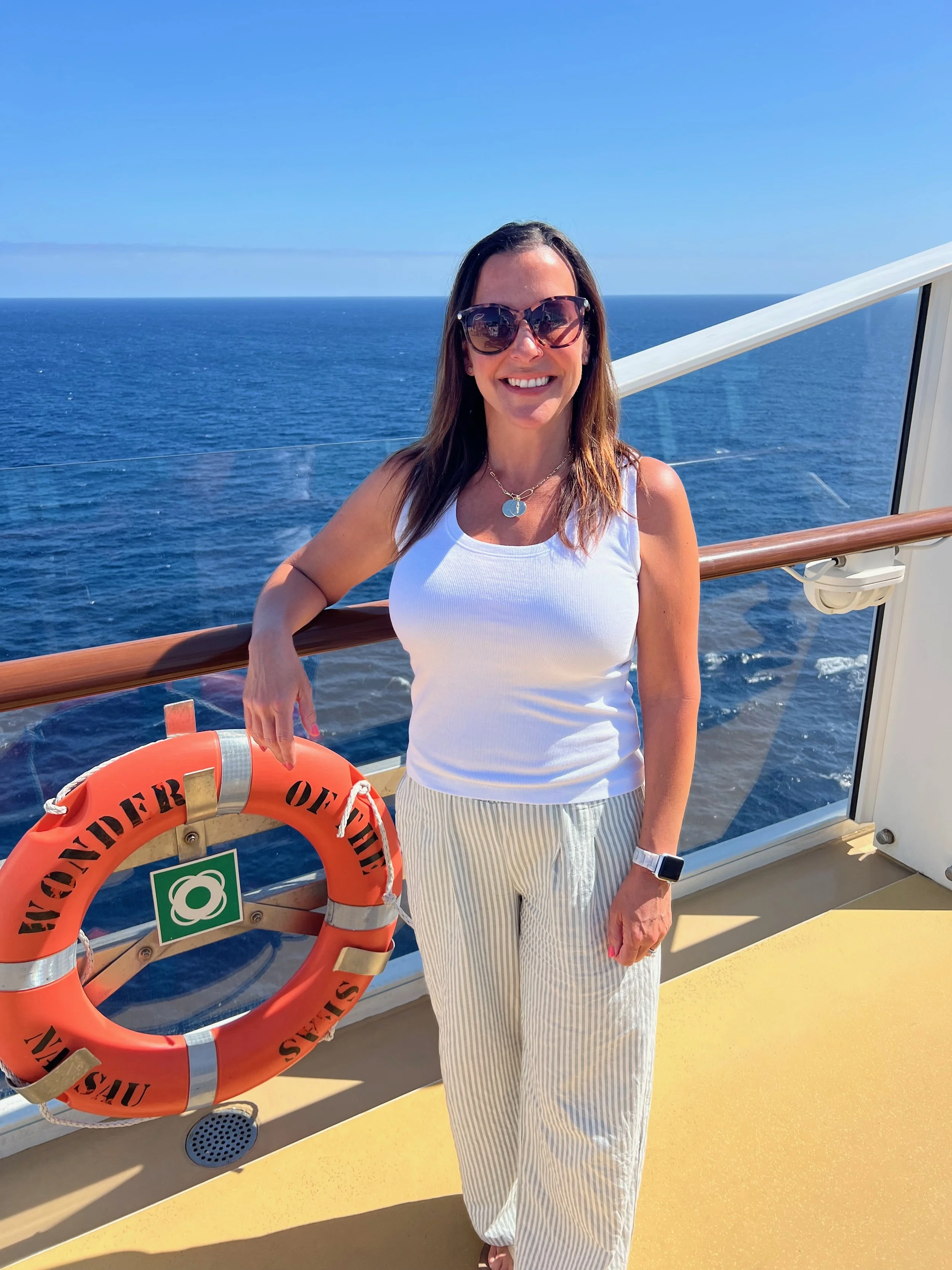 A woman in sunglasses and a white tank top stands on a cruise ship deck with ocean in the background. She is smiling and leaning on a lifebuoy.