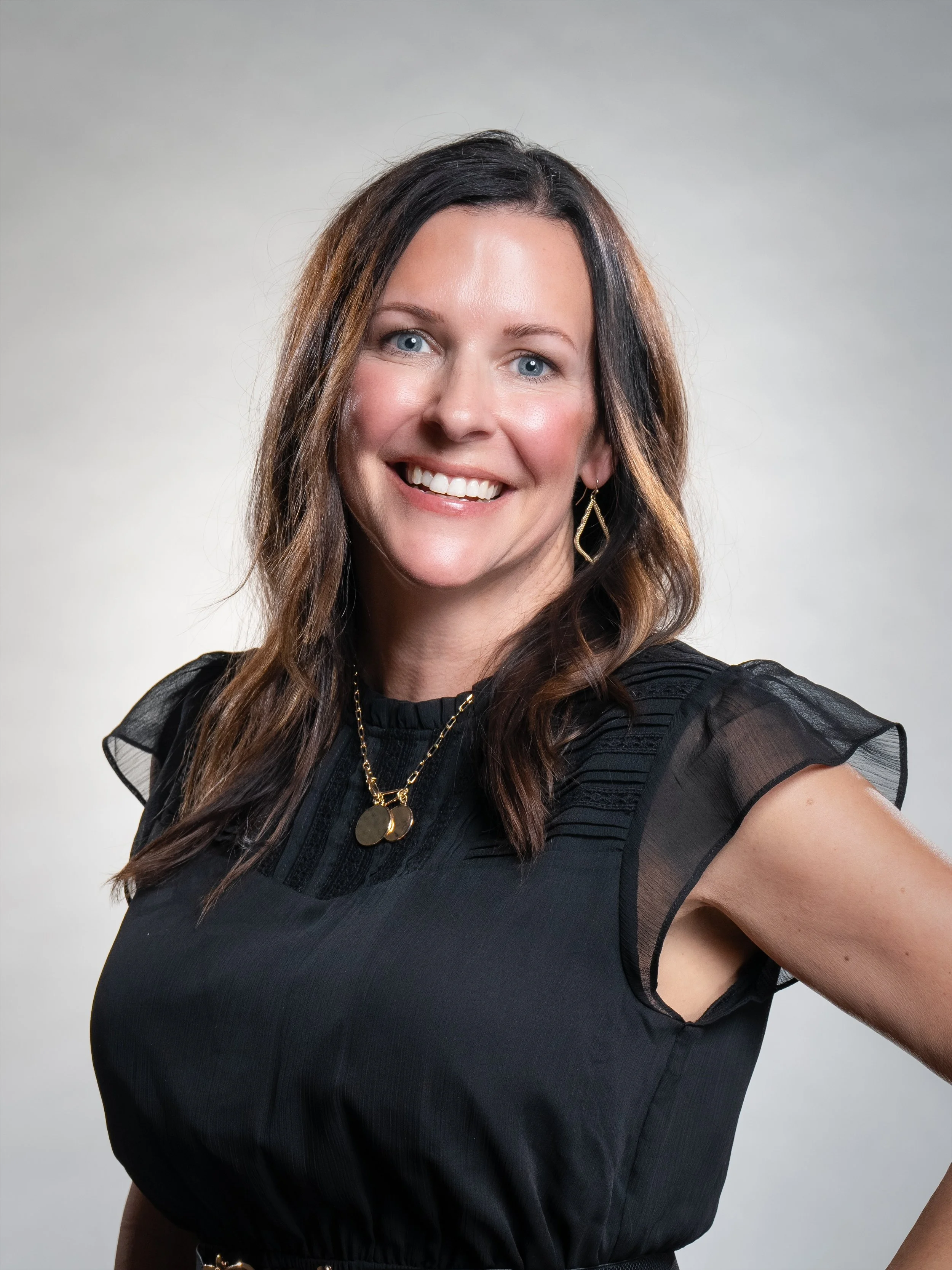 A woman with long brown hair and blue eyes, smiling, wearing a black dress with sheer sleeves, gold jewelry including a necklace and earrings, against a neutral background.