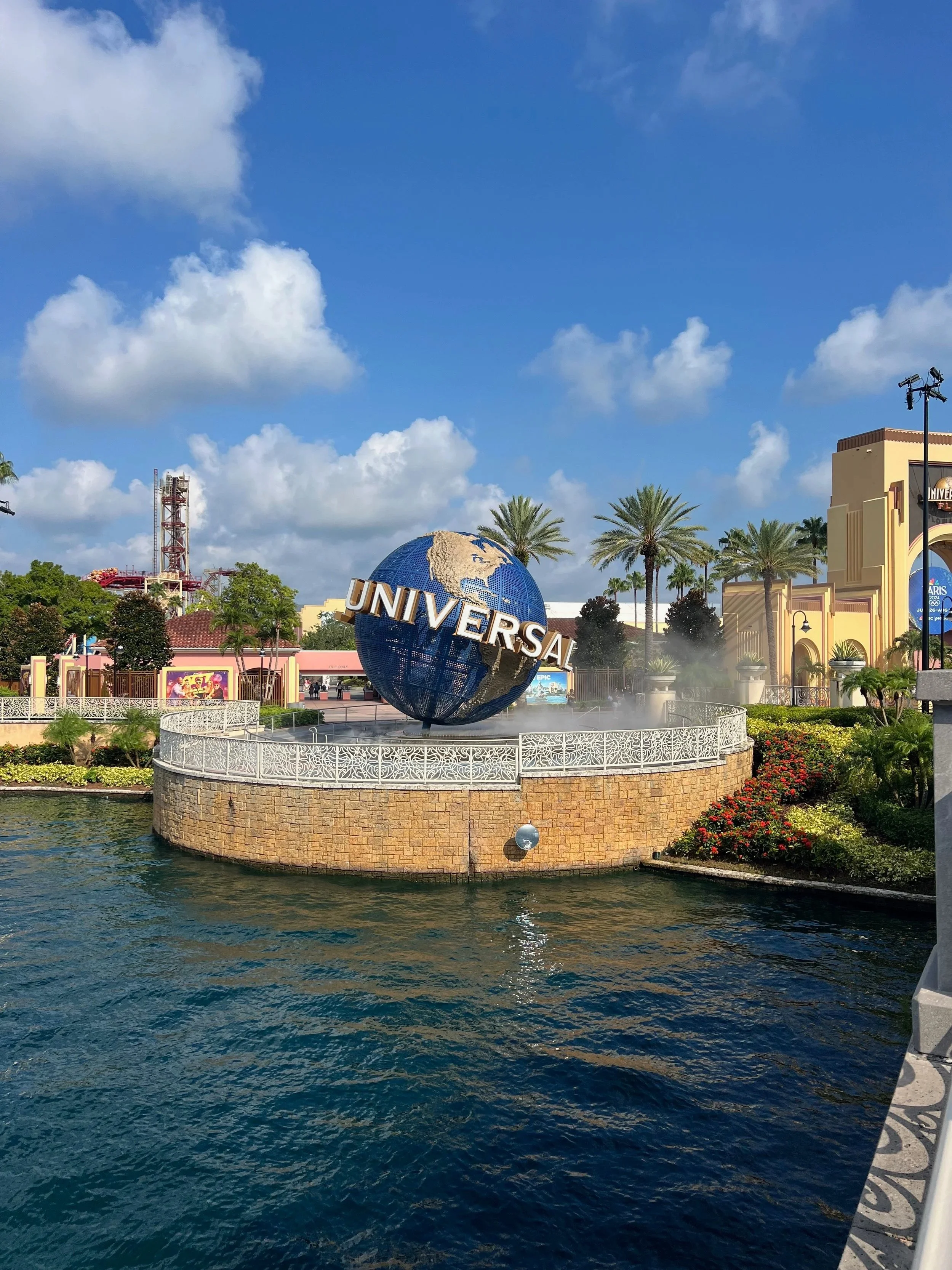 Universal Studios logo with a globe fountain, surrounded by water, palm trees, and theme park buildings.