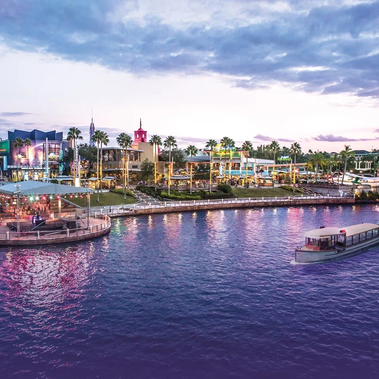 A waterfront scene at dusk with a boat on calm water, illuminated buildings, palm trees, and a cloudy sky.