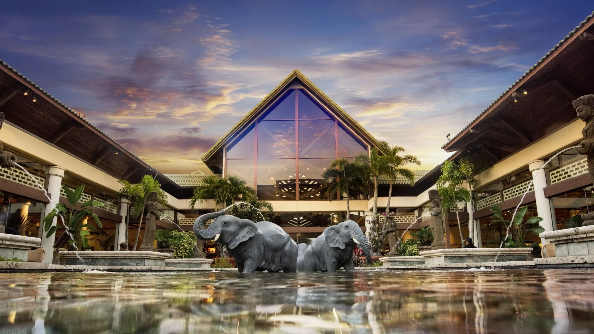 A courtyard with two elephant sculptures in a fountain, surrounded by tropical plants and statues, in front of a modern building with large glass windows and a triangular roof against a sunset sky.