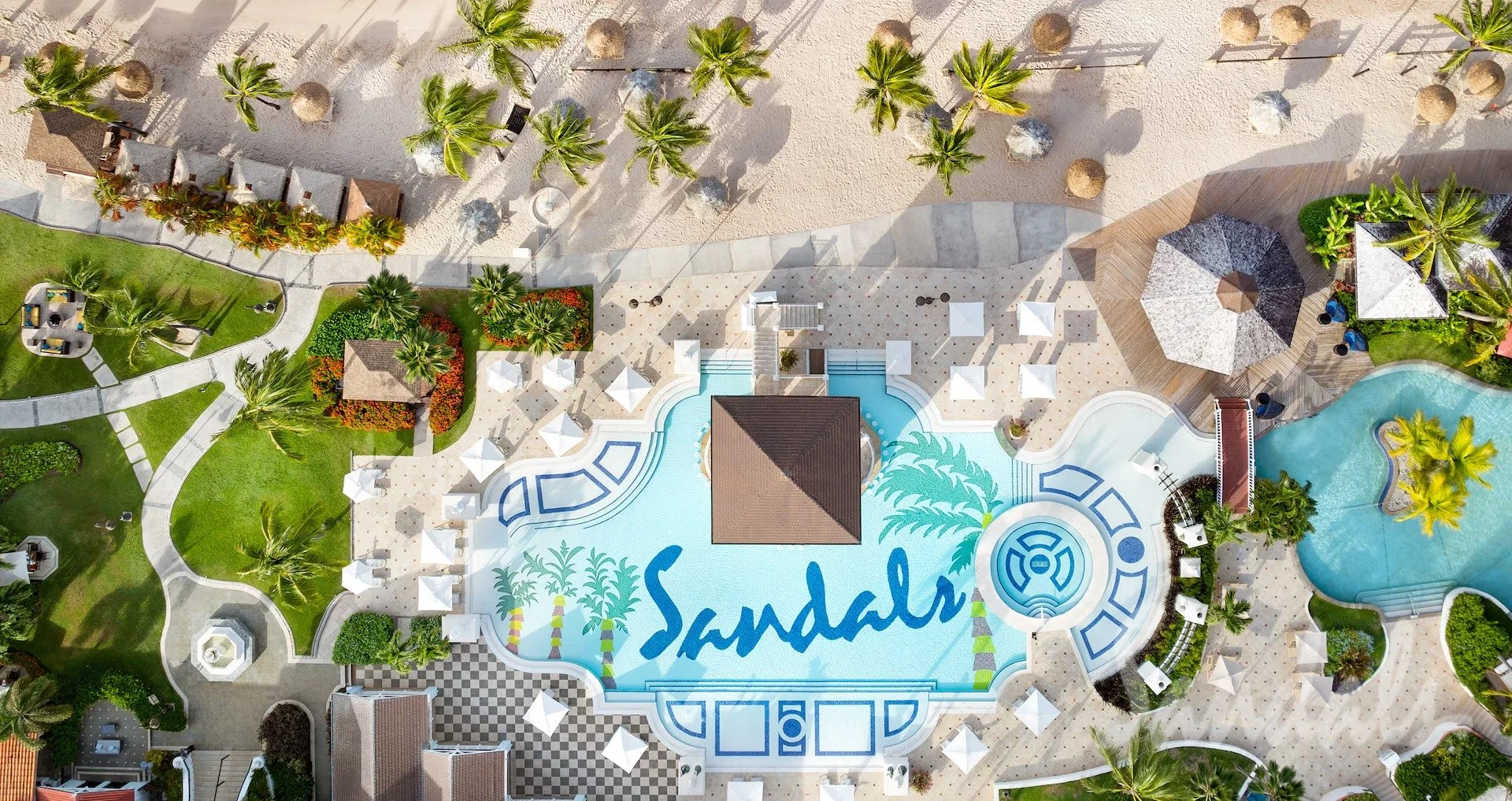 Aerial view of a resort pool area with a large pool featuring the word "Sandals" and palm trees, surrounded by greenery, lounge chairs, umbrellas, and pathways, with a sandy beach and additional structures nearby.