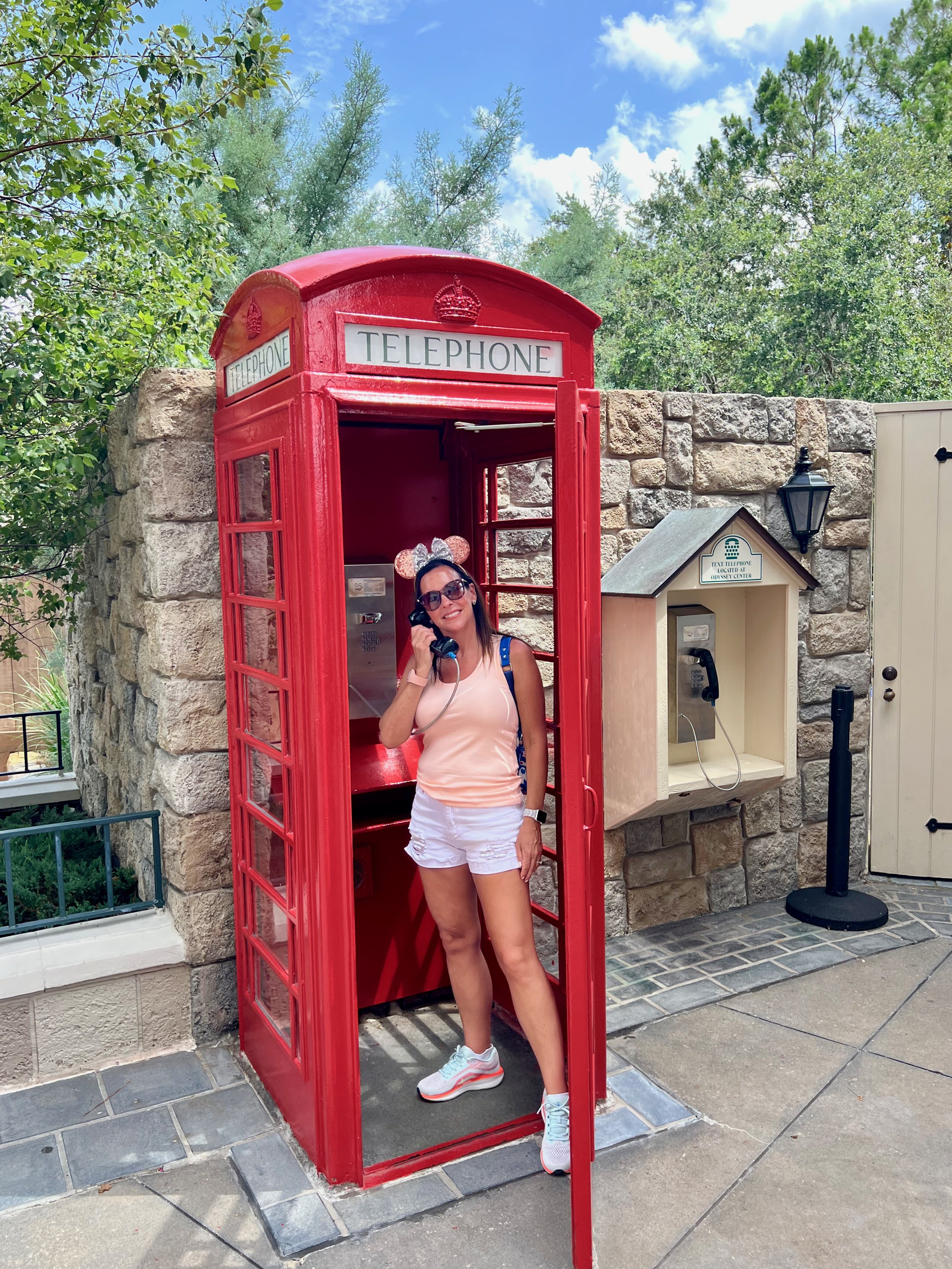 A woman wearing sunglasses, a pink tank top, white shorts, and sneakers, standing inside a red vintage telephone booth outdoors. She is talking on the phone and smiling.