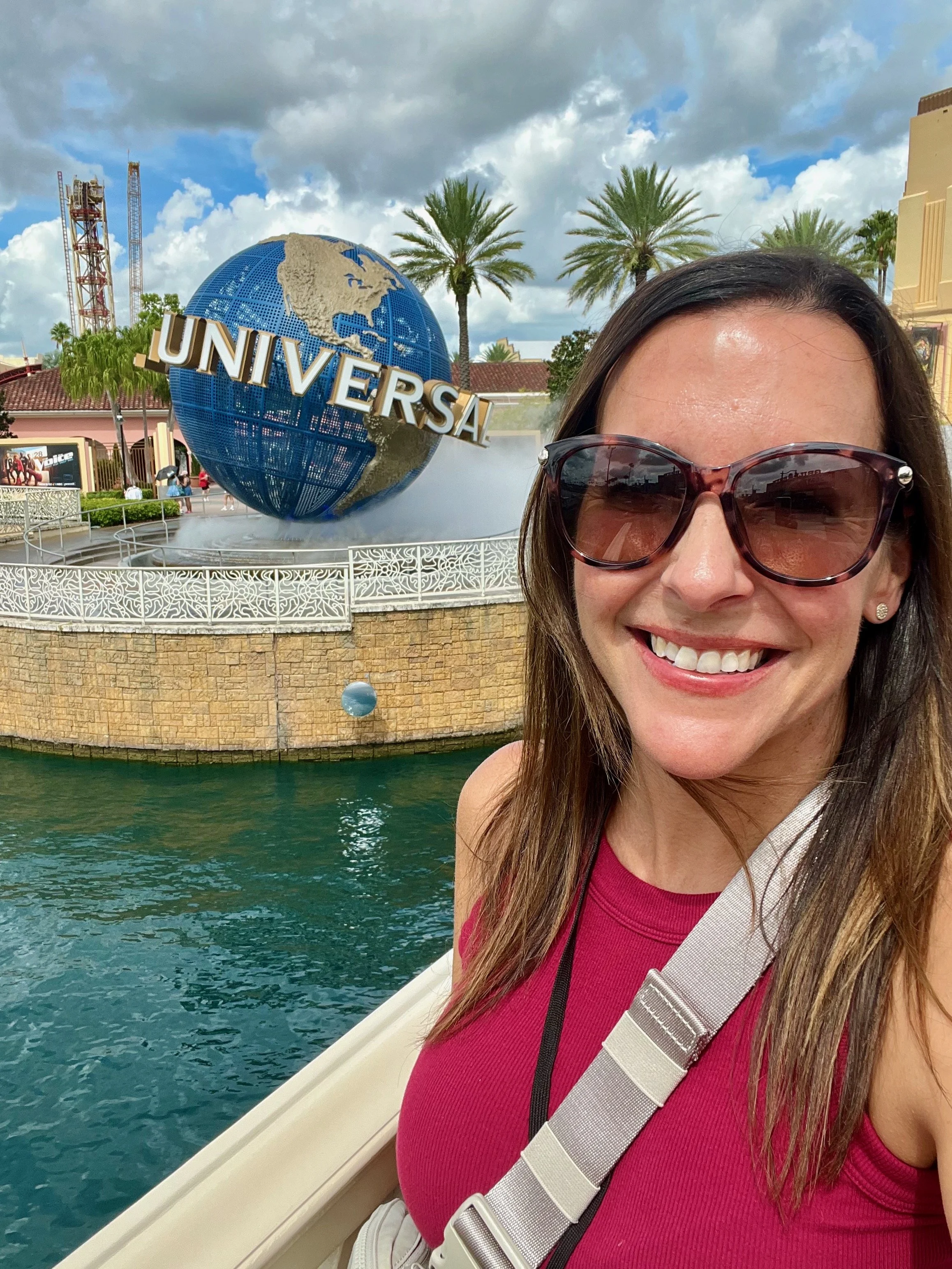 A woman smiling in front of the Universal Studios globe at Universal Orlando Resort, wearing sunglasses and a red top.