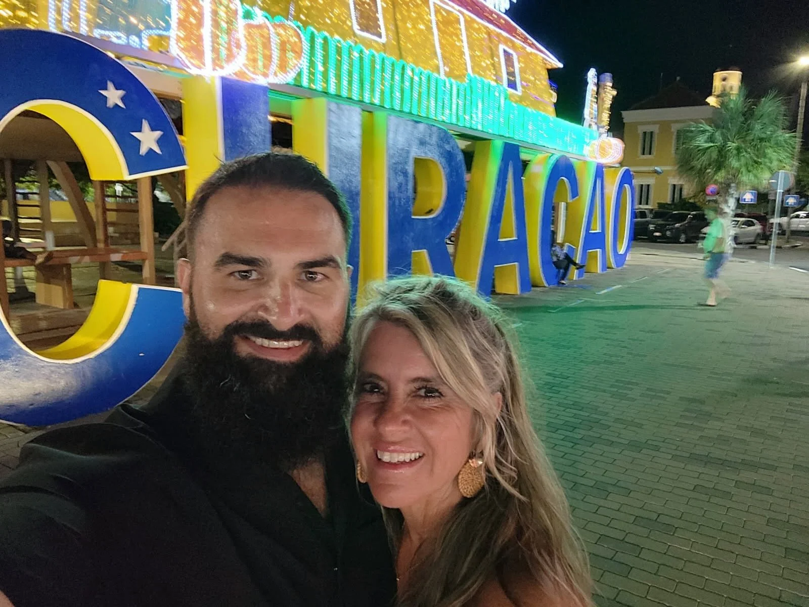 A selfie of a smiling man and woman in front of colorful, illuminated 'CURACAO' sign at night.