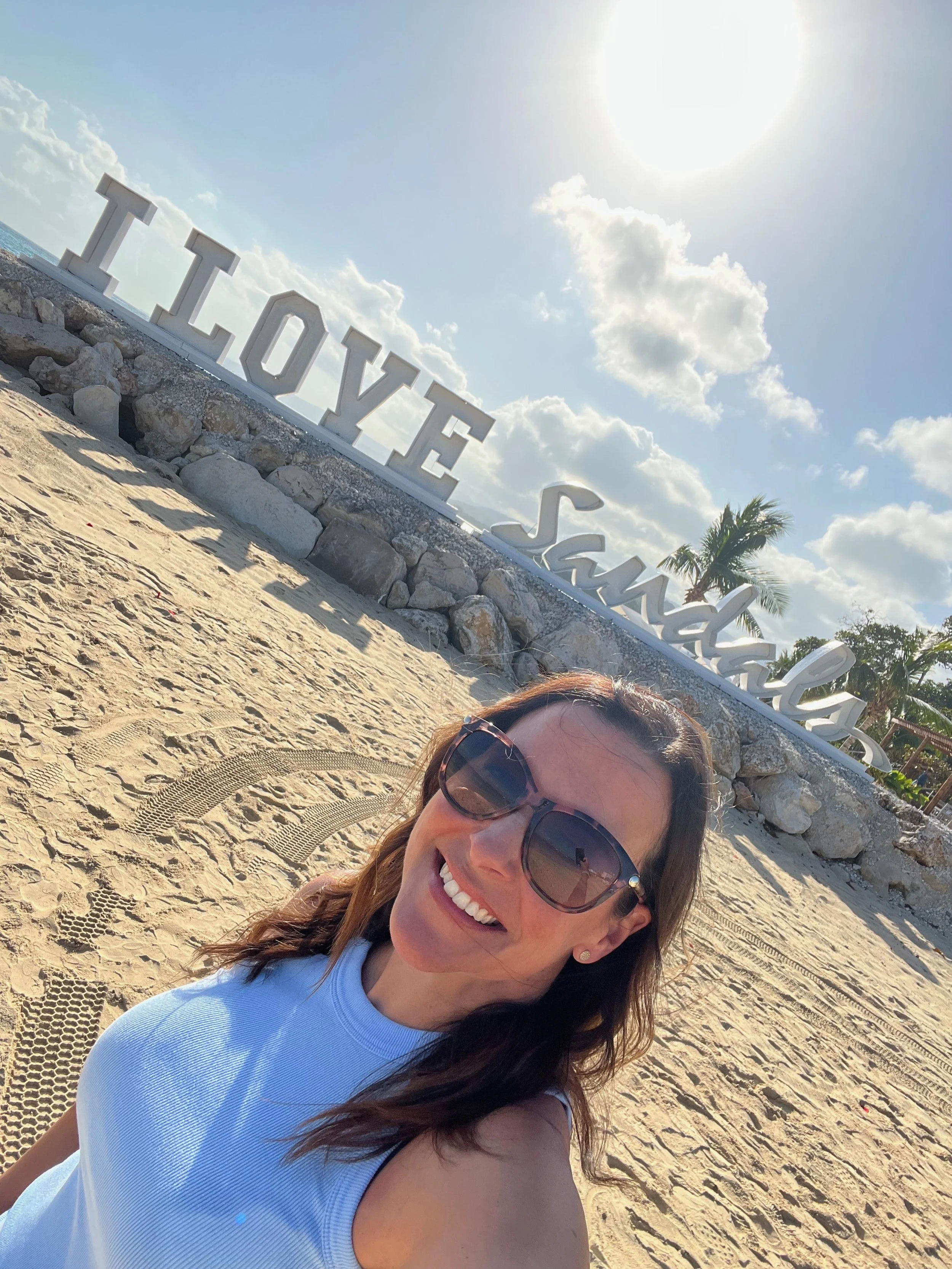 Woman smiling on a sandy beach with large white 'I LOVE SANDALS' sign and palm trees in the background under a sunny sky.
