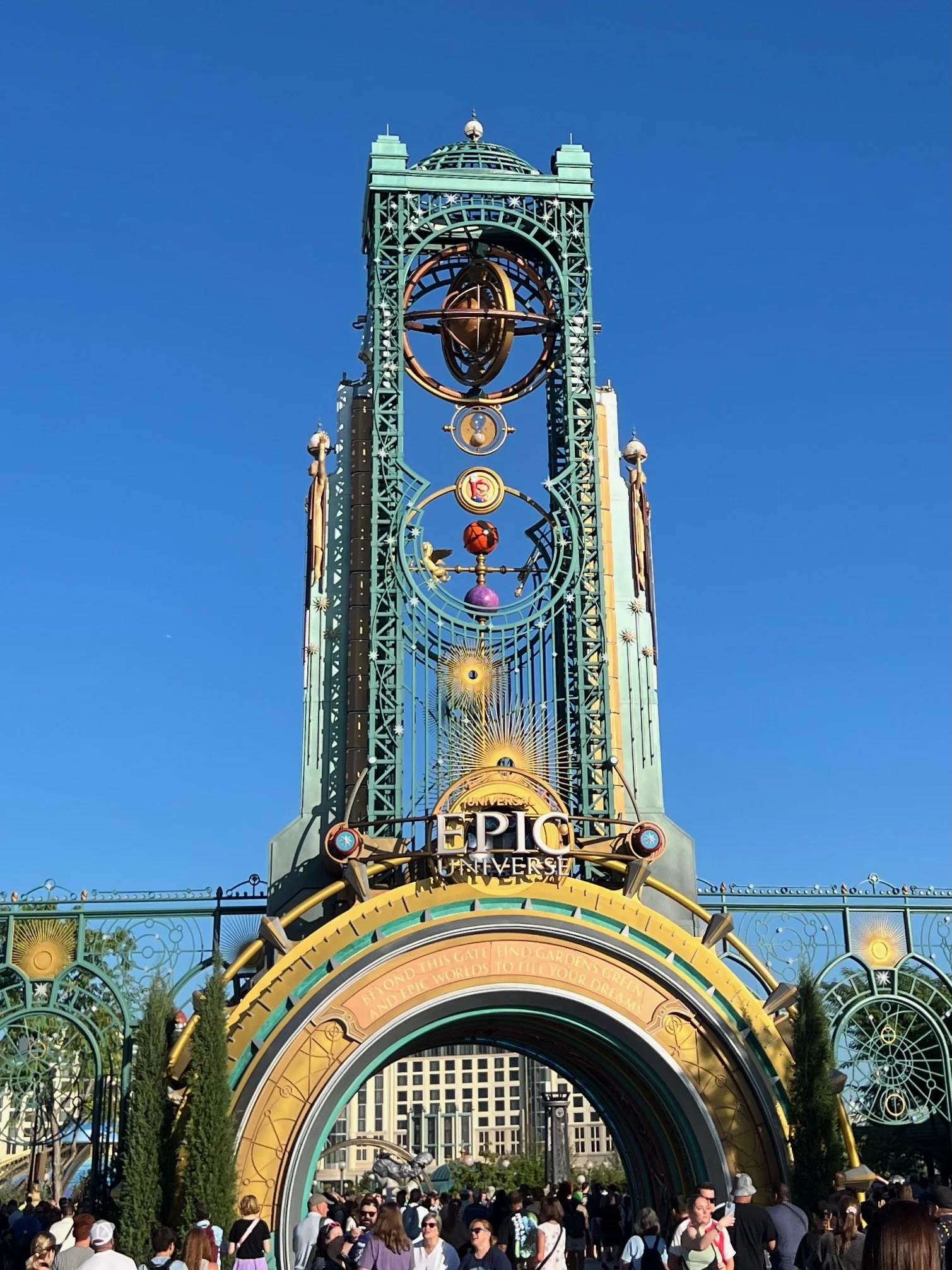 Entrance gate at Universal's Epic Universe theme park with a tall decorative clock tower and a crowd of visitors.
