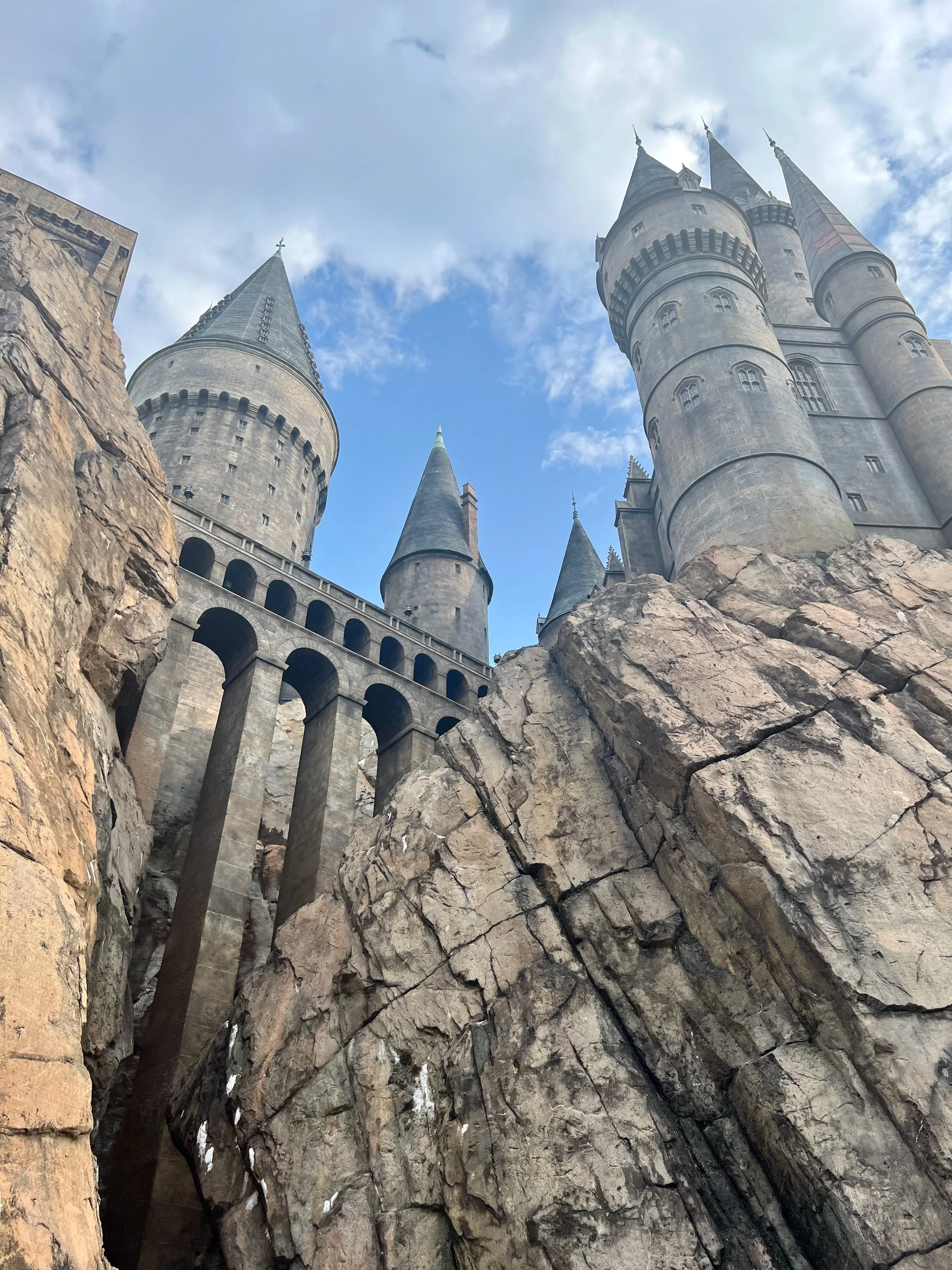 View of Hogwarts with multiple towers and turrets built into a rocky landscape under a partly cloudy sky.