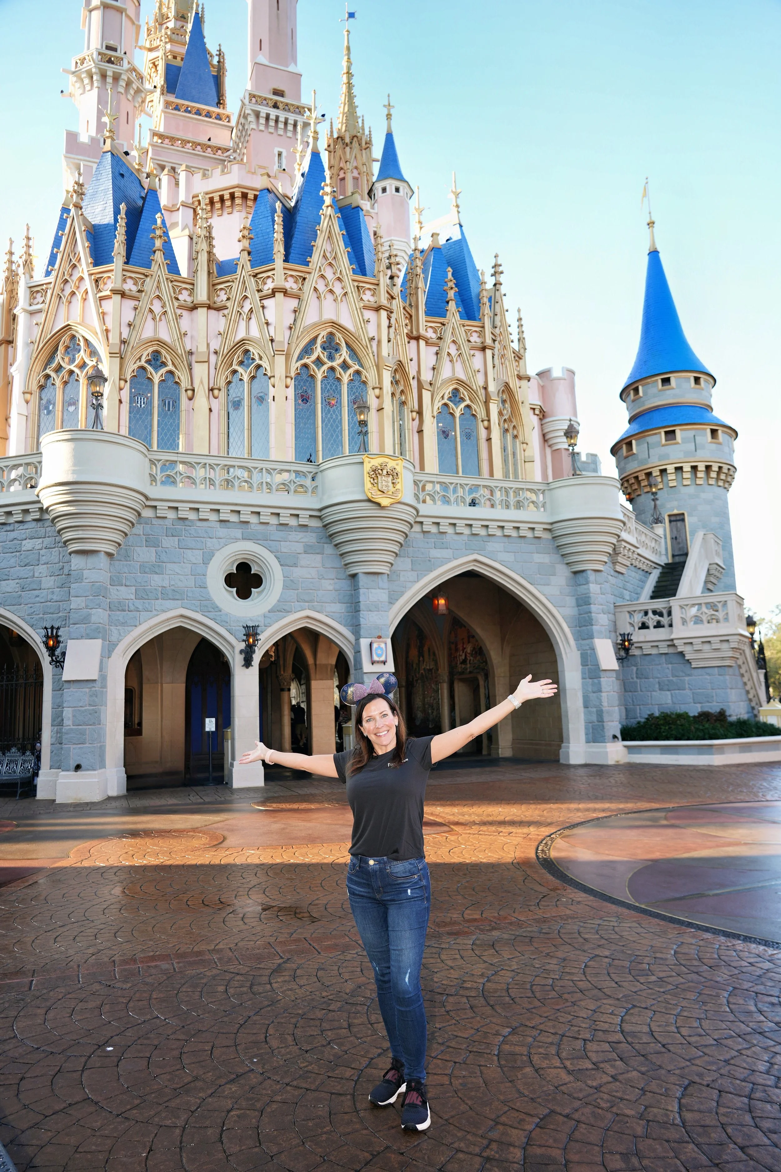 A woman with Mickey Mouse ears poses happily with arms open in front of Cinderella Castle at Disney World.