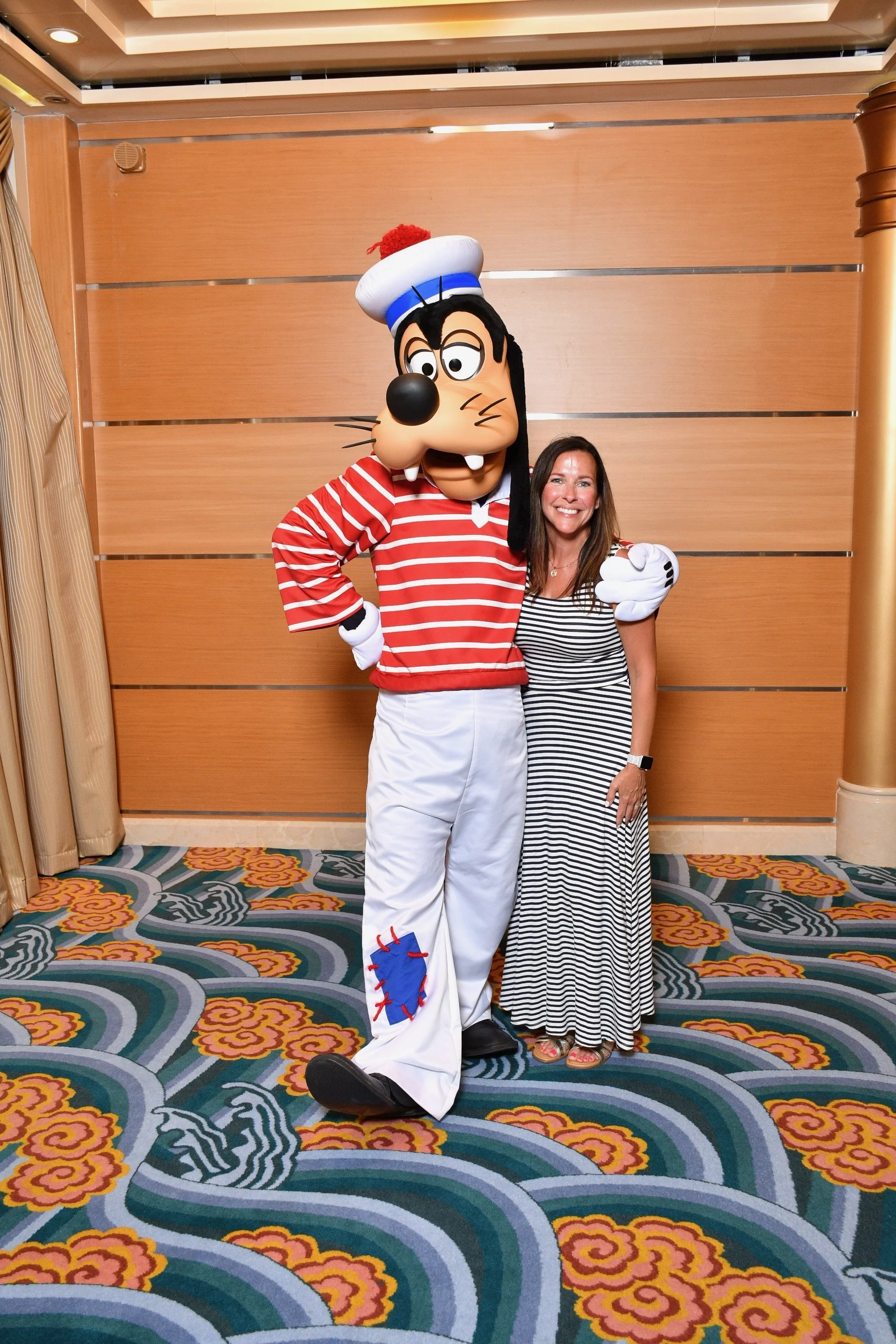 A woman in a striped dress standing next to a person dressed as the cartoon character Goofy, who is wearing a red and white striped shirt, white pants with a blue patch, and a sailor hat, in a decorated indoor space.