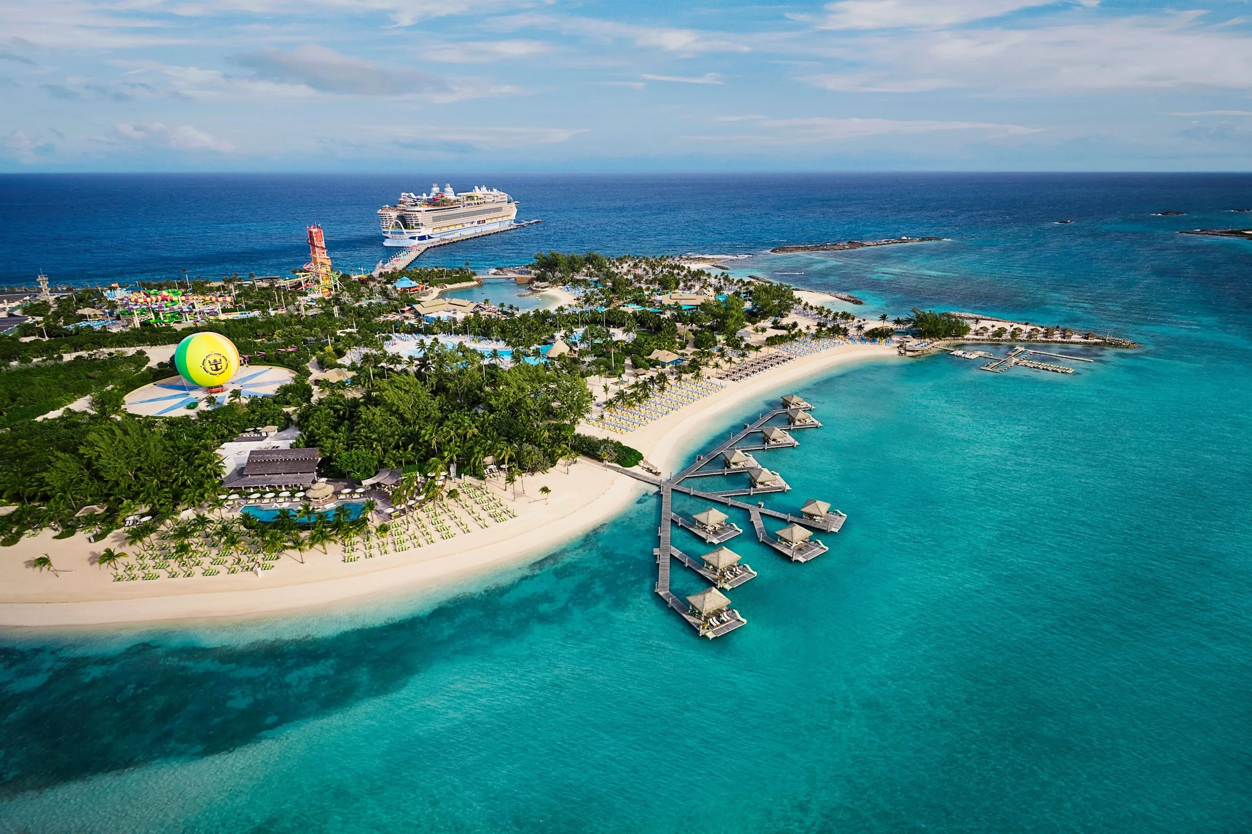 Aerial view of a tropical island resort with white sandy beaches, overwater bungalows, a large cruise ship in the ocean, a hot air balloon, and a water park with a tower, surrounded by turquoise water and lush green trees.