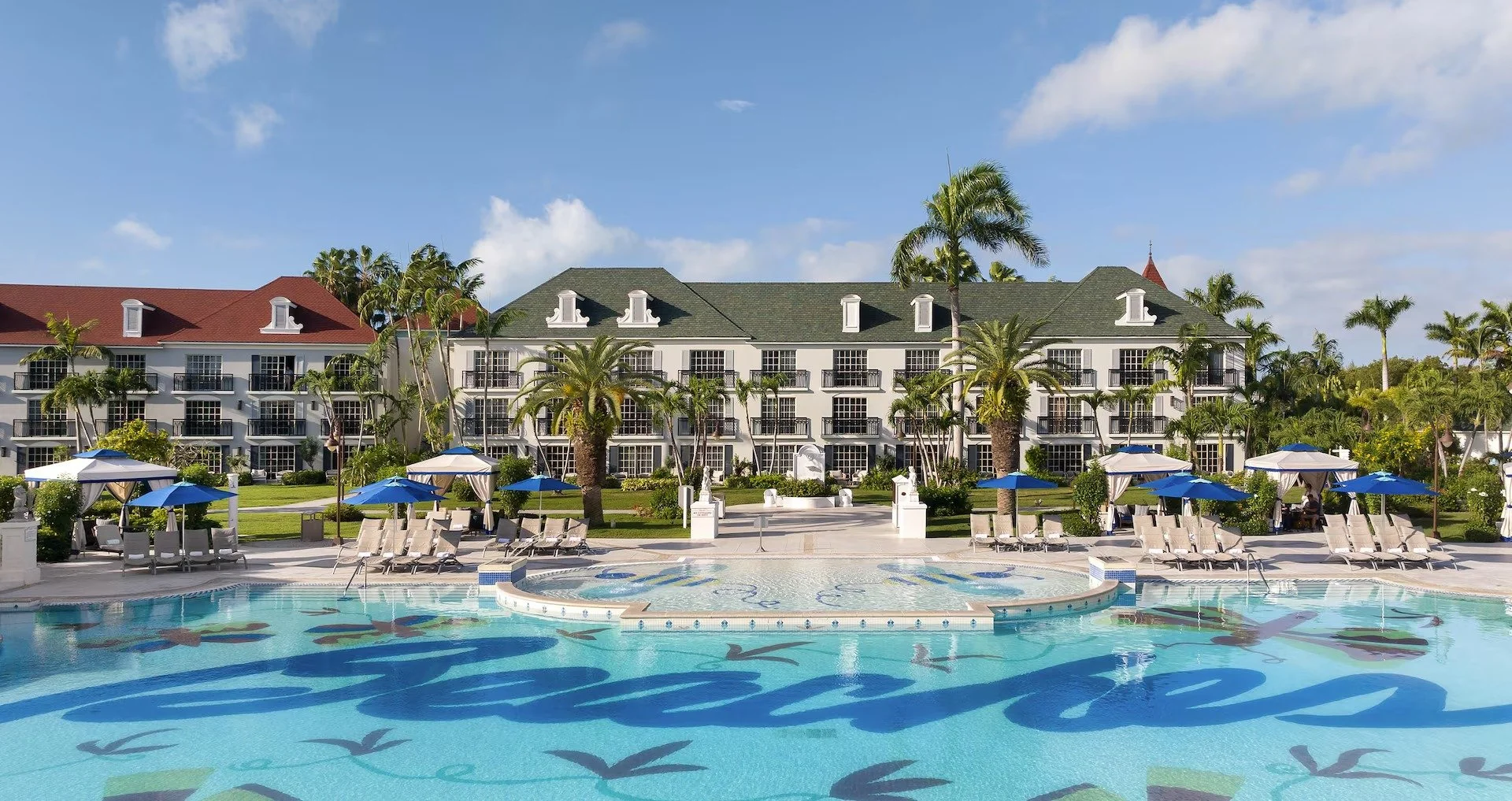 Resort pool area with lounge chairs, umbrellas, and a hotel building in the background, surrounded by palm trees under a blue sky.