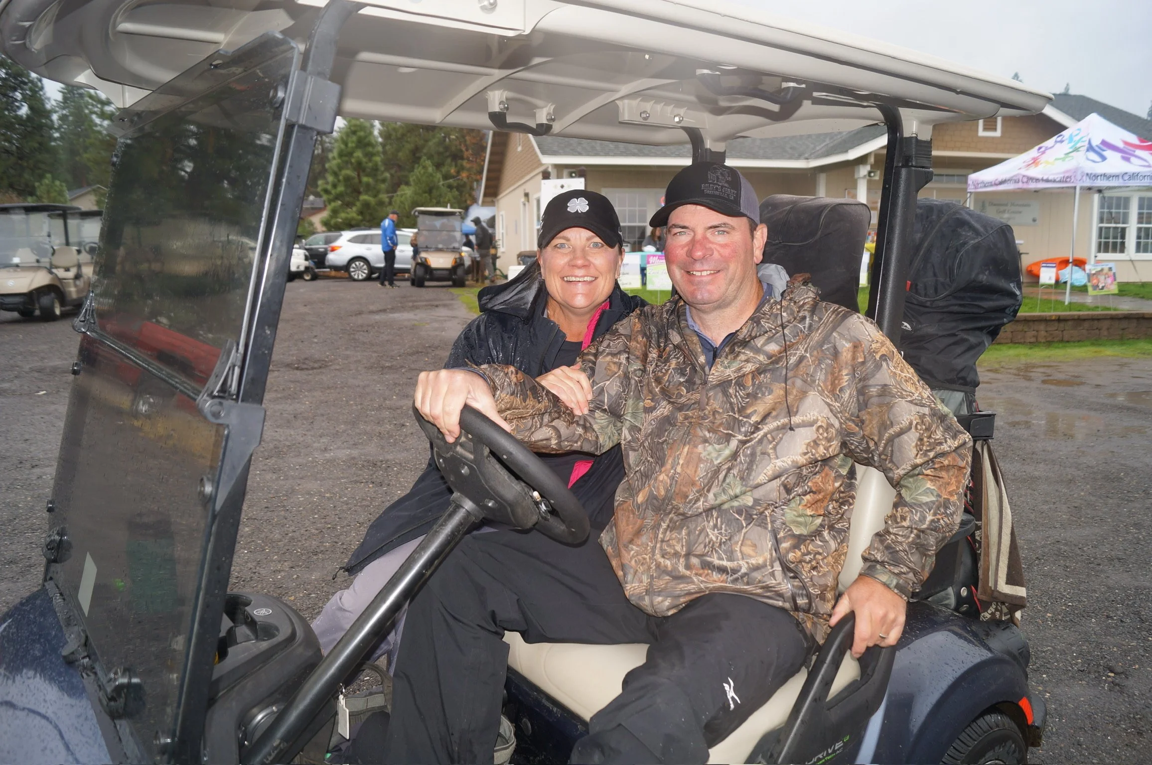A smiling couple sitting in a golf cart outdoors, with trees and buildings in the background during a light rain.