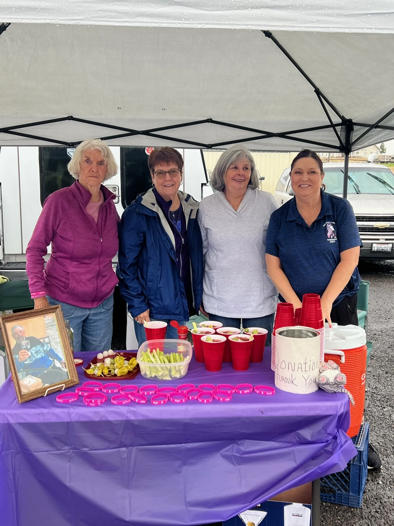 Four women standing behind a table with red cups and plastic containers, under a canopy at an outdoor event. One woman is wearing a purple jacket, others in casual clothing. There is a framed photo of a man in a racing suit on the table, and a note r