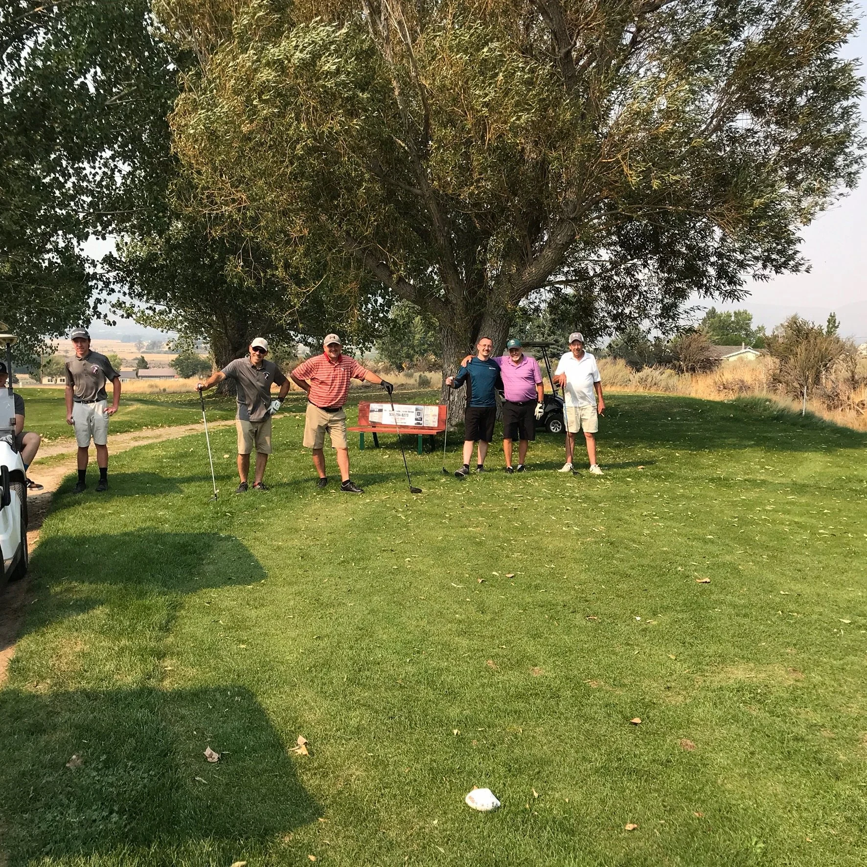 A group of six people standing on a golf course under a large tree, with a golf cart in the background and a golf ball on the grass in the foreground.