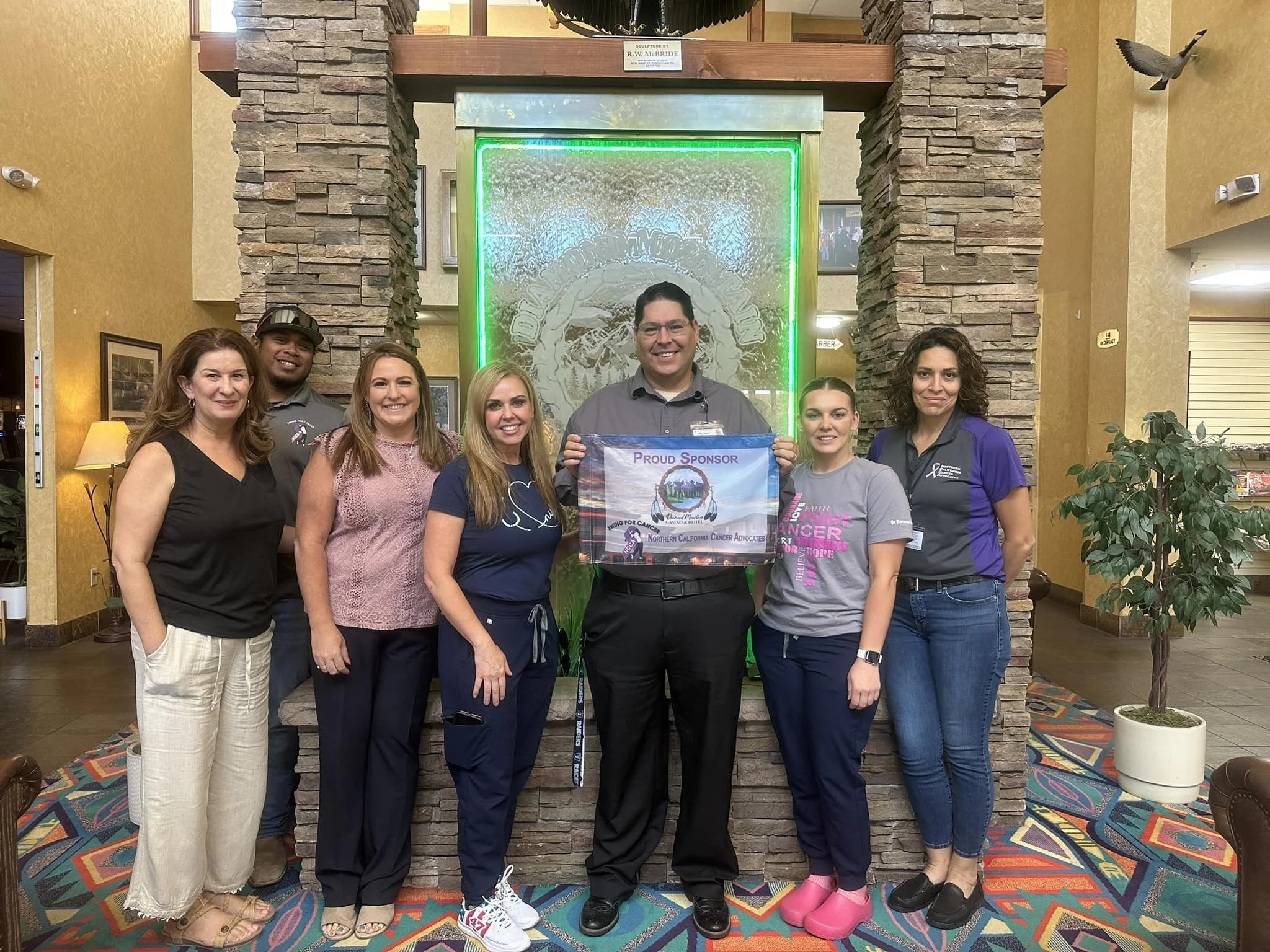 Group of seven people standing in front of a leaf design and a green neon sign, holding a flag that reads 'Proud Sponsor' for a cancer organization, in an indoor setting with a colorful carpet and a potted plant.