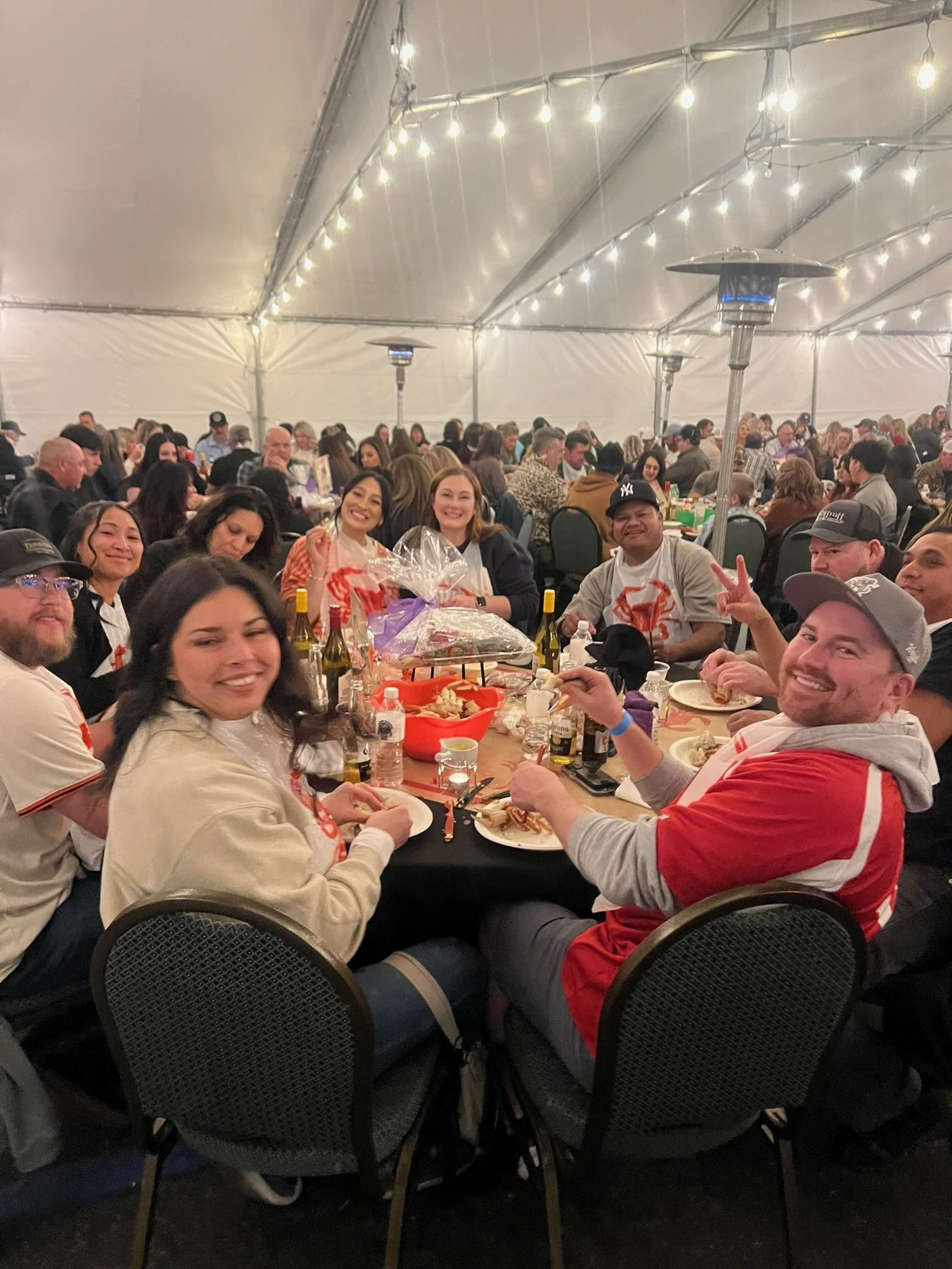 A group of people gathered around a long table inside a large tent, enjoying a meal together at a social event.