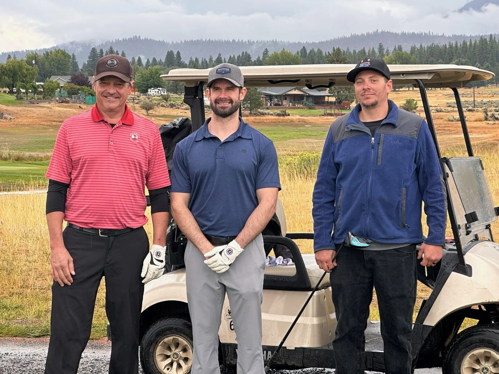 Three men standing next to a golf cart outdoors on a golf course, with trees and mountains in the background.