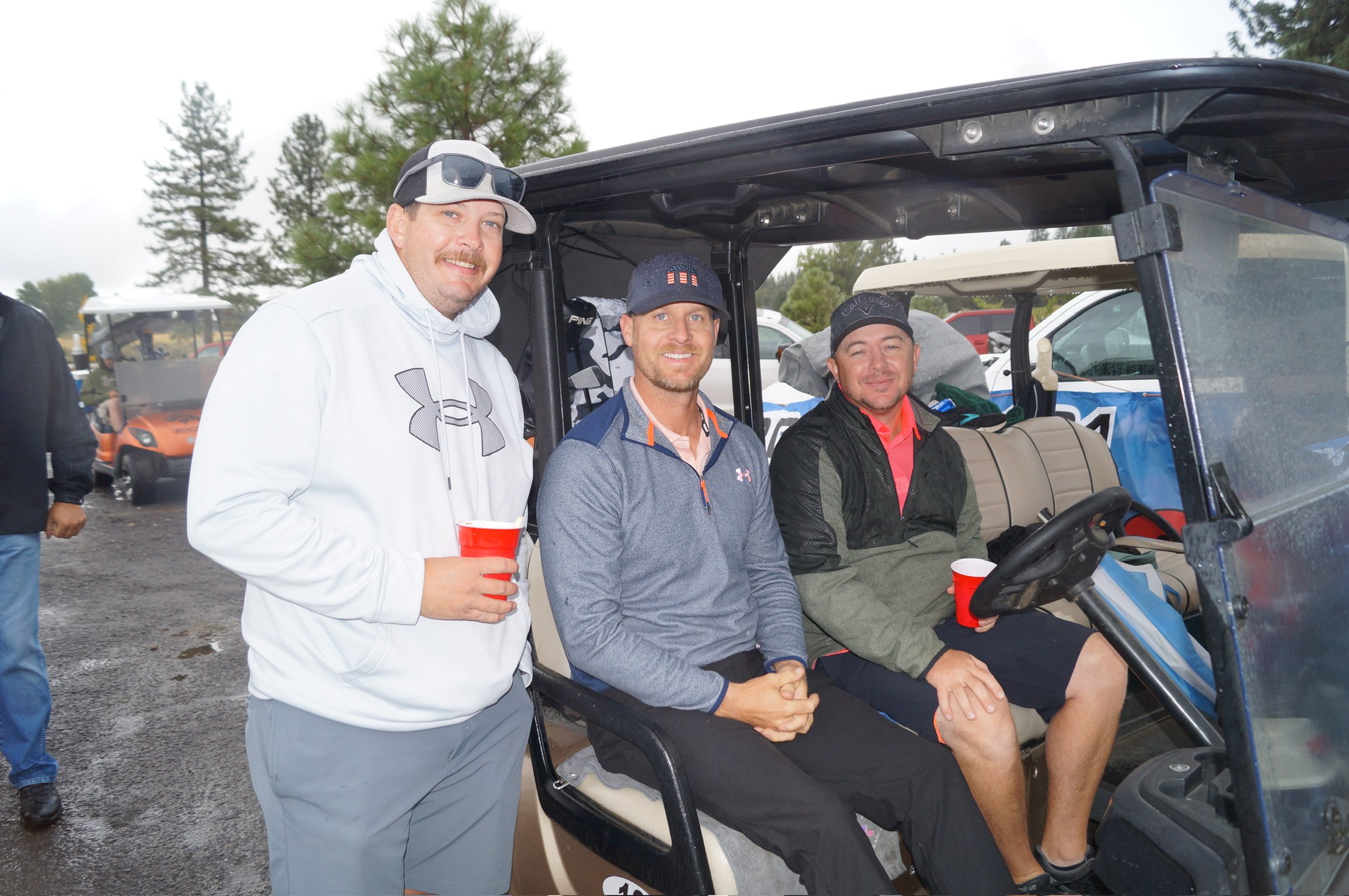 Three men in casual clothing sitting and standing next to a golf cart, with trees and parked vehicles in the background. One man is holding a red cup and smiling, while the other two are seated in the golf cart, smiling at the camera. The scene appea