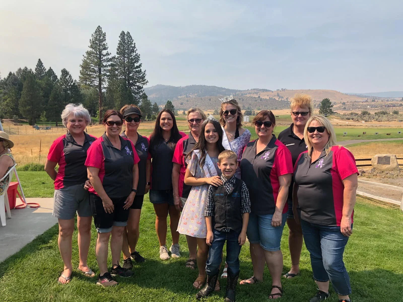 A group of eleven people, including children and adults, posing outdoors in a rural landscape with rolling hills and trees in the background. The people are smiling and some are wearing matching pink and gray shirts, possibly for a team or event.