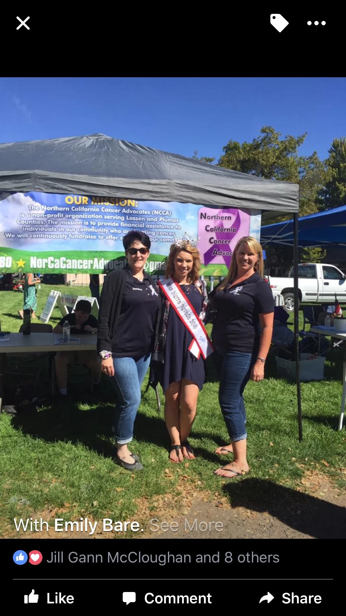 Three women standing in front of a tent with a banner that reads 'Our Mission: The Northern California Cancer Advocates.' One woman wears a sash that says 'Miss California Jr.' The scene is outdoors with a grassy area and trees in the background.