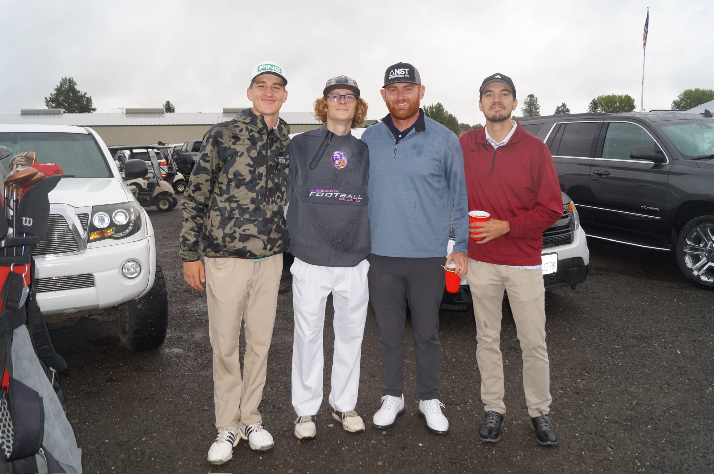 Four young men standing together outdoors in a parking lot, with cars and golf carts behind them, under a cloudy sky, one holding a red cup, dressed casually and smiling.