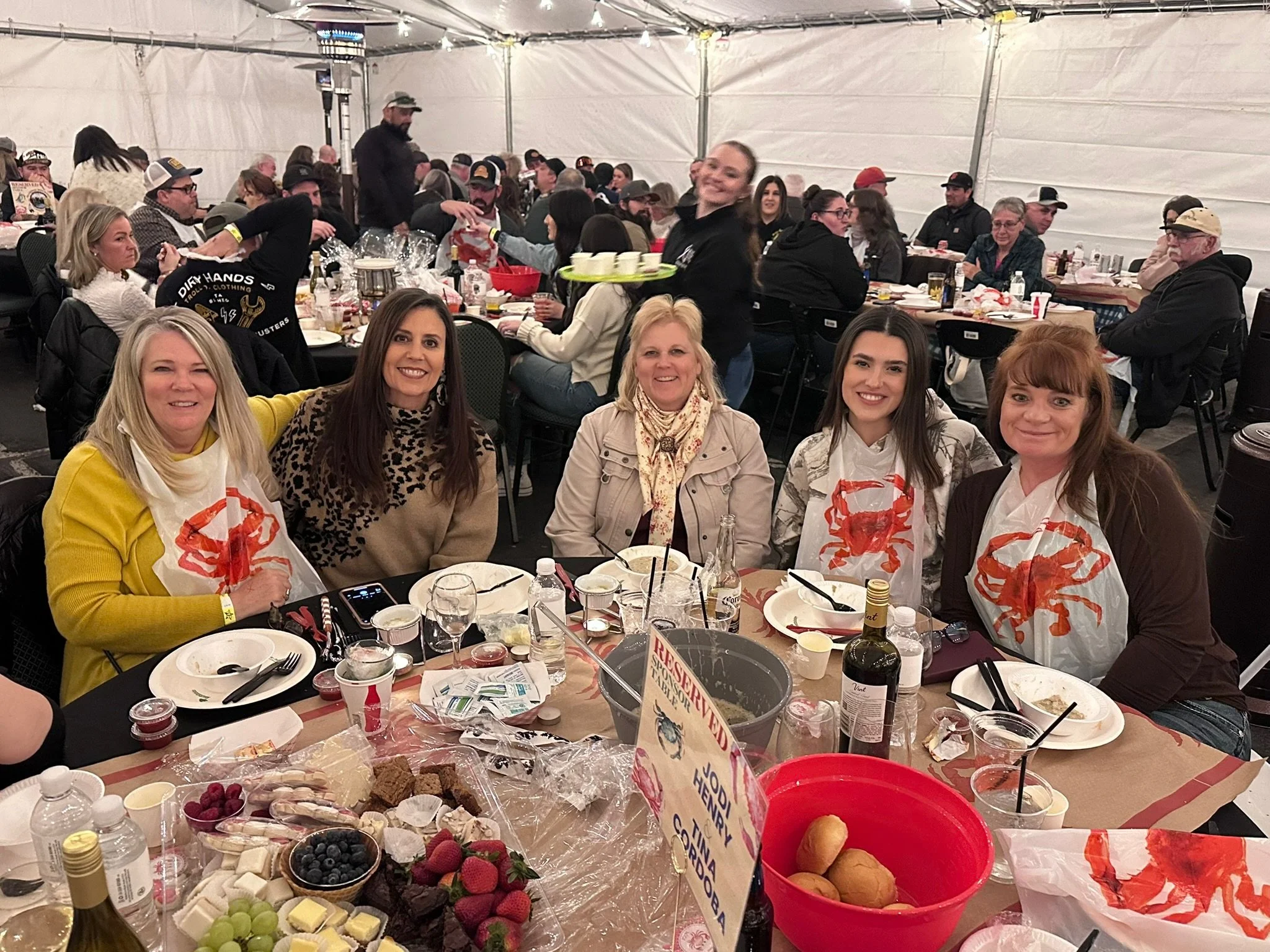 A group of people at a banquet table inside a tent, smiling and enjoying food. There are various dishes, fruit, and drinks on the table, and some guests are wearing crab-themed clothing. A woman in the center is smiling at the camera and a server is holding a tray with cups.