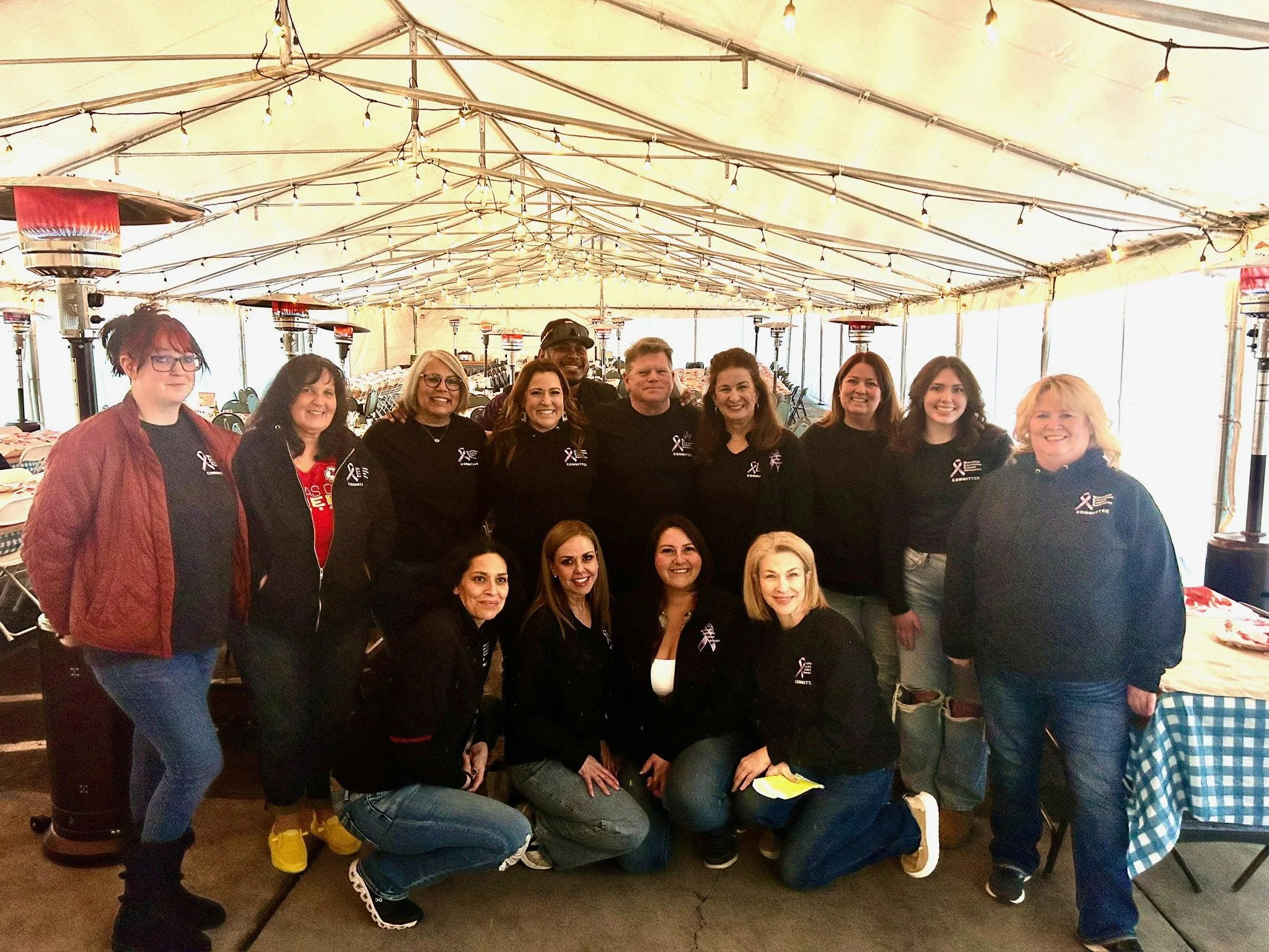 Group of women and one man posing together inside a large tent decorated with string lights. Several people are smiling, wearing casual clothing including black shirts with a ribbon logo, and standing close to each other.