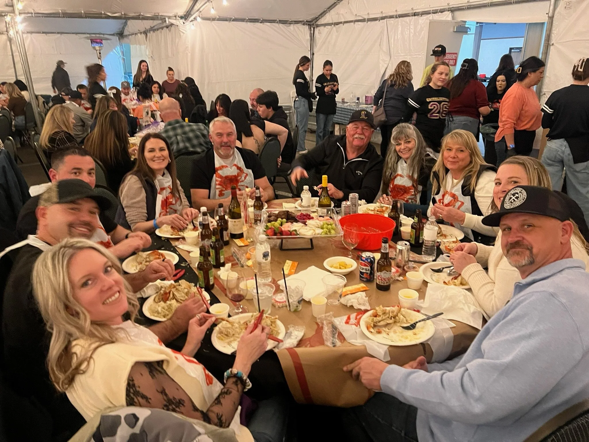 People sitting and eating at a large gathering or party inside a tent, with tables filled with food and drinks.