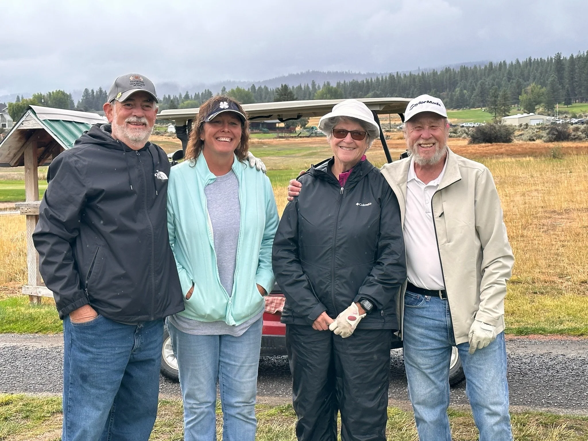 Four people standing together outdoors on a cloudy day, smiling. Two men and two women, dressed casually, with a golf cart and a golf course in the background.