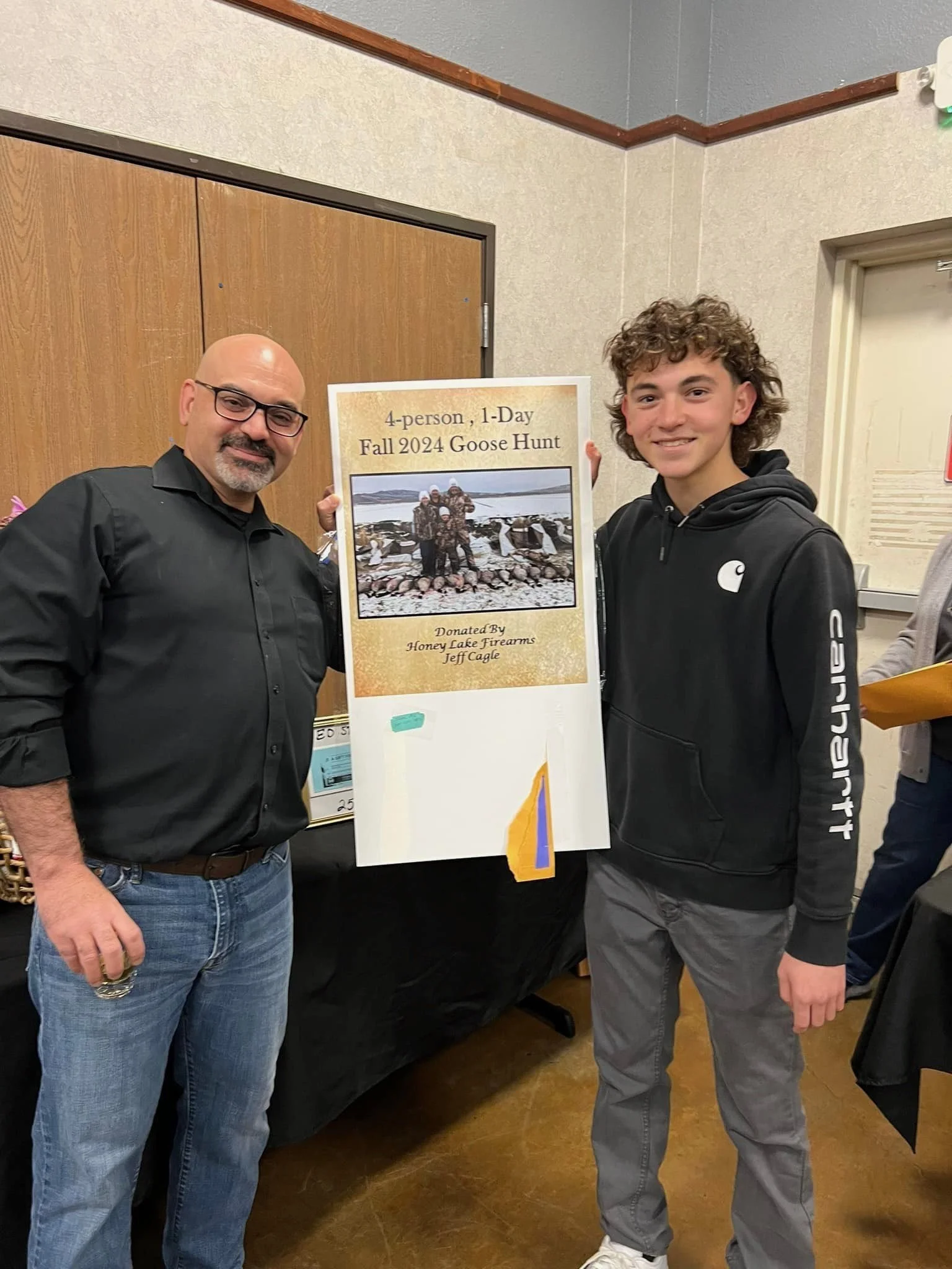 Two men are standing indoors near a poster about a fall 2024 goose hunt. The man on the left is wearing glasses, a black shirt, and jeans. The man on the right is younger, wearing a black hooded jacket and gray pants. They are both smiling.