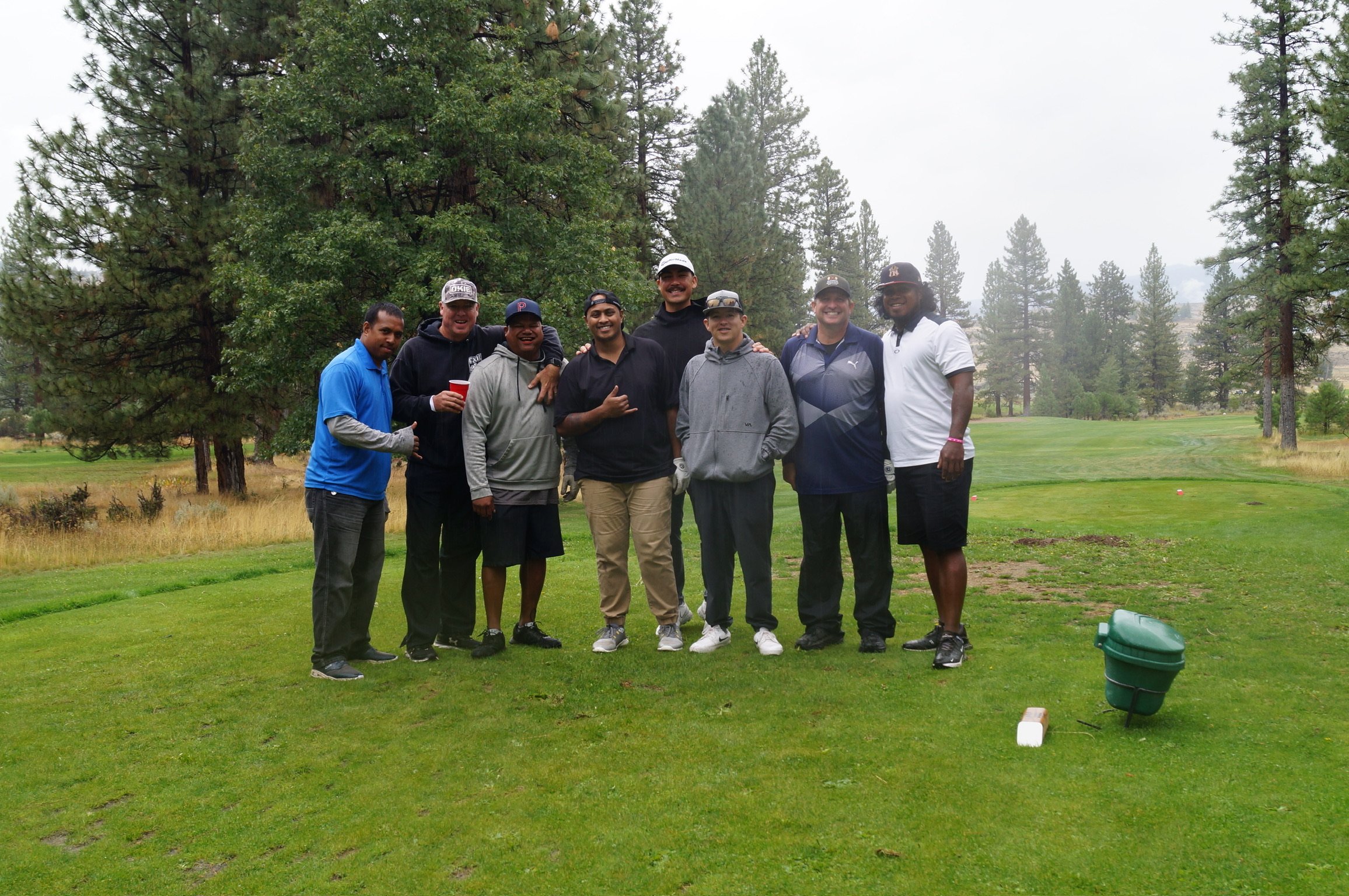 A group of eight men standing together on a golf course, smiling for a photo amid trees and greenery.