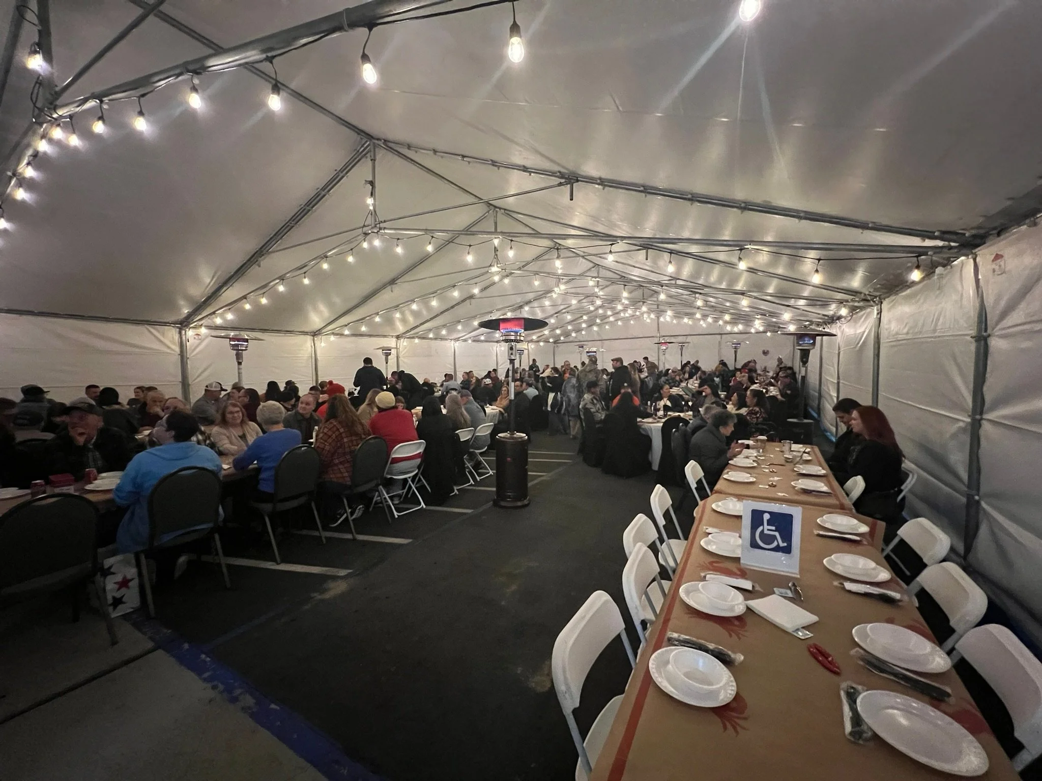 Indoor event tent with string lights, filled with many people sitting at tables. The tables are set with plates and utensils, and there are patio heaters providing warmth. A sign indicates accessible seating.