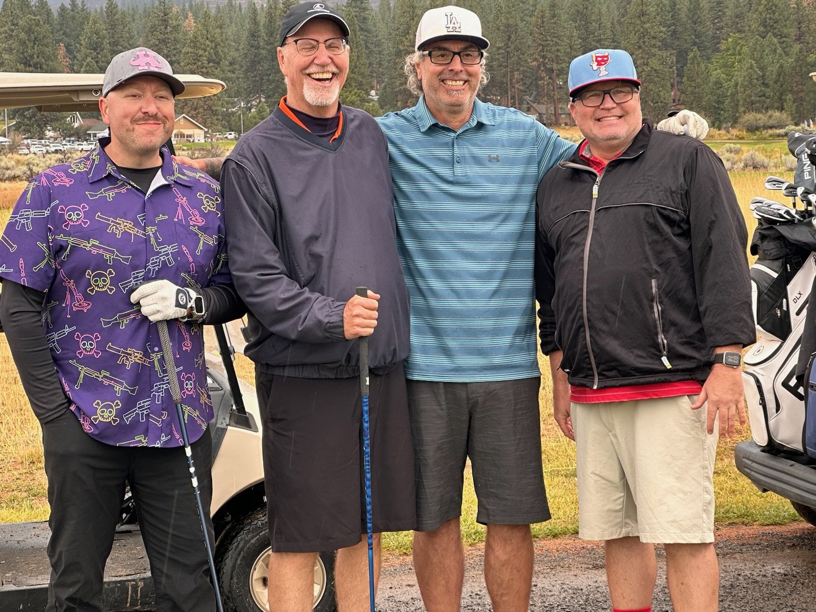 Four men dressed in golf attire standing together on a golf course with golf carts and trees in the background, smiling.
